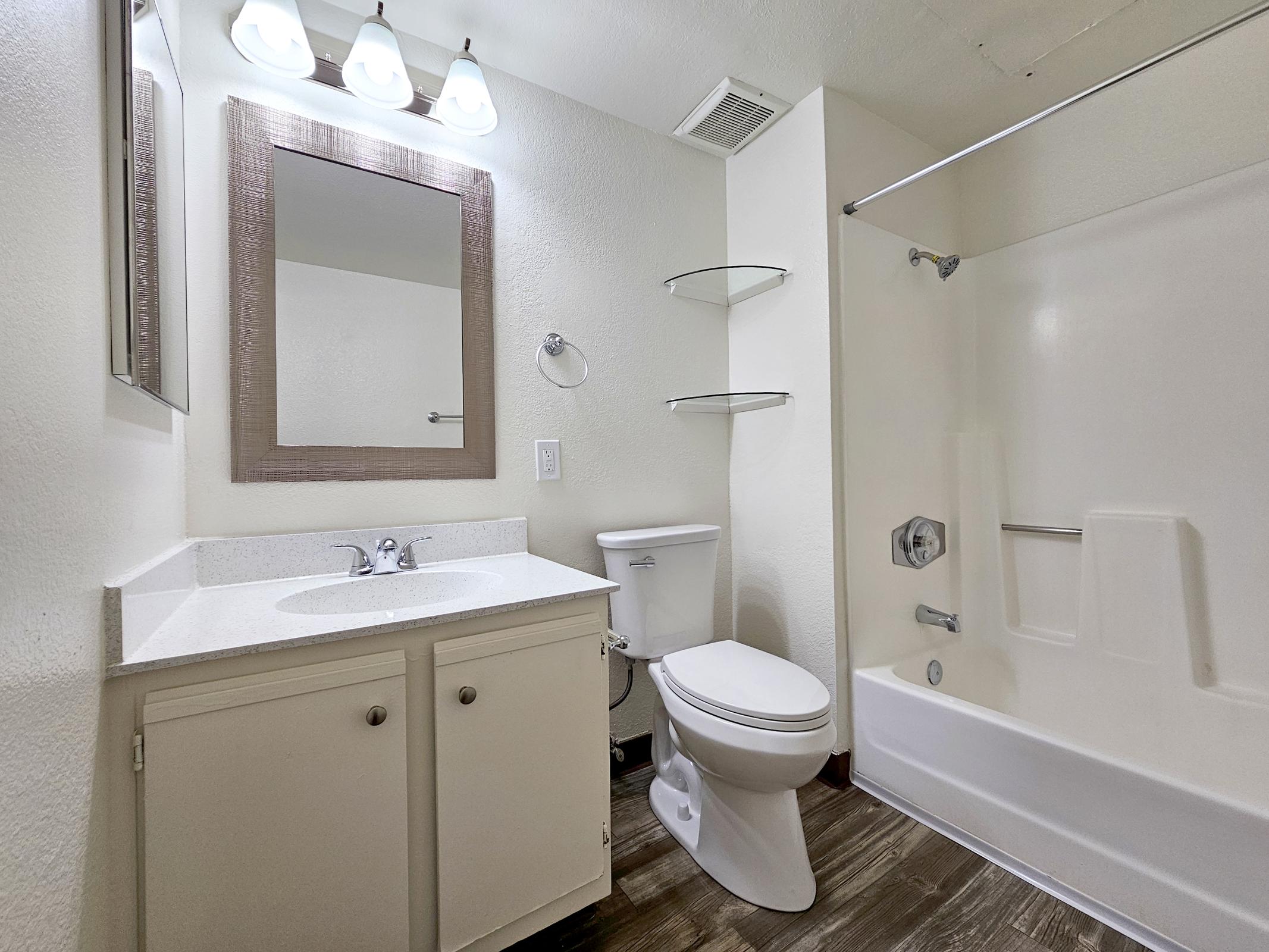 A clean and modern bathroom featuring a white shower/tub combo, a toilet, and a vanity with a sink. The space is illuminated by three light fixtures above a mirror. There are shelves for storage and the flooring is a wood-like material, giving a contemporary look to the room.