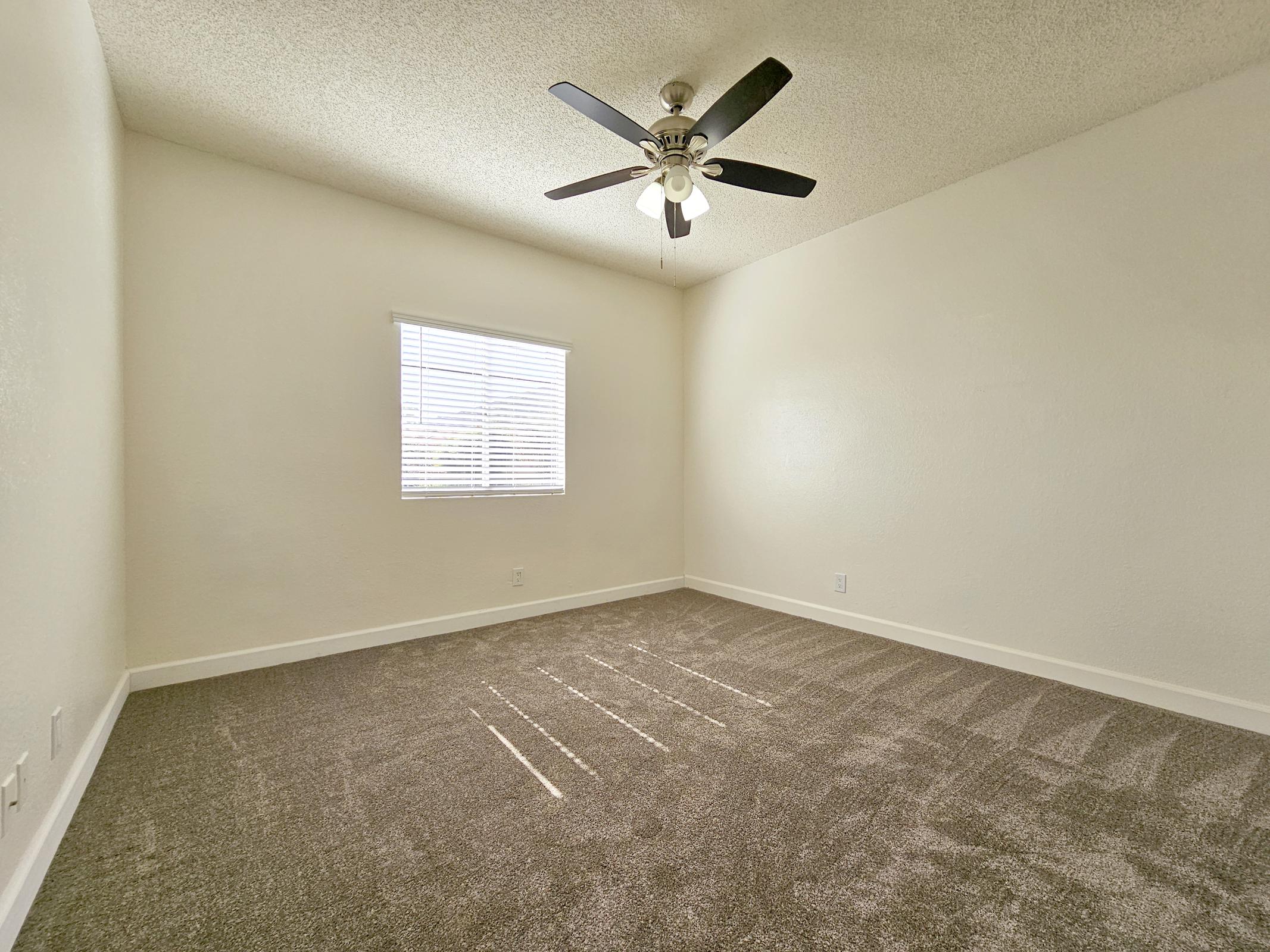 A spacious, empty room with light beige walls and a ceiling fan. The floor is covered with plush, light brown carpet. A window with white blinds allows natural light to enter, casting soft shadows on the floor. The room has a clean, minimalist aesthetic, ideal for personal customization.