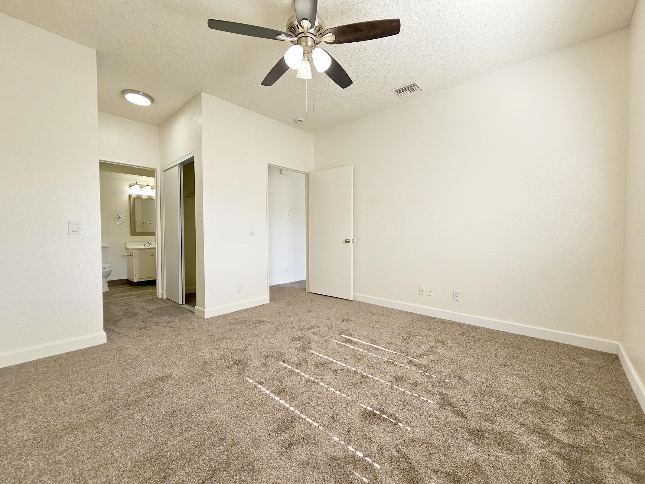 A spacious, unfurnished room with light-colored walls and carpeted floor. A ceiling fan is mounted on the ceiling. To the left, there is an open doorway leading to a bathroom, and a closet is visible on the right. Natural light streams in from a window, illuminating the space.