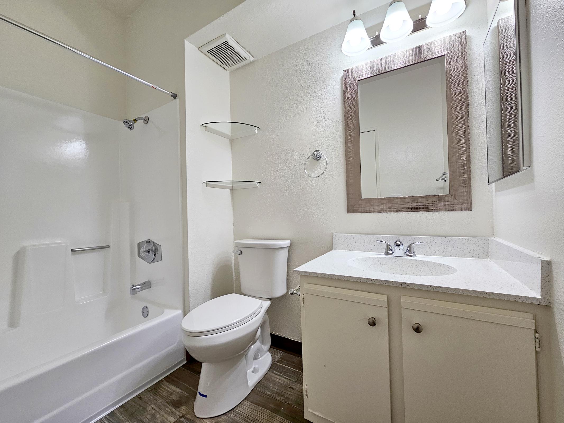 A clean and modern bathroom featuring a white bathtub with a shower curtain, a toilet, a vanity with a sink, and a large mirror above it. The walls are painted a light color, and there are shelves for storage. Natural light illuminates the space from a ceiling light fixture.