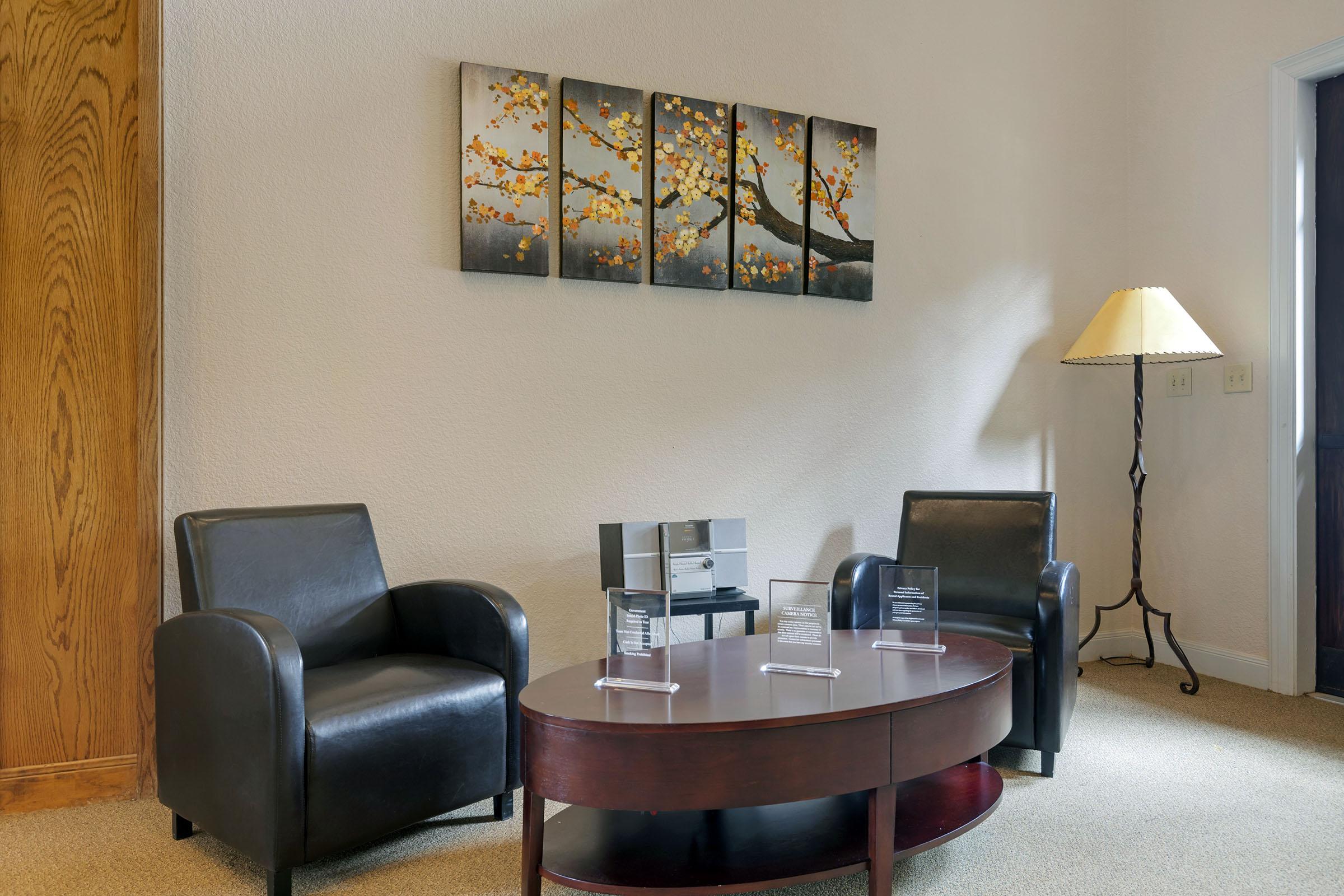 A cozy waiting area featuring two black chairs, a round wooden coffee table, and an elegant lamp. On the wall hangs a series of four canvases depicting a tree with orange leaves. There are also a couple of clear display stands on the table. The room has a warm and inviting ambiance.