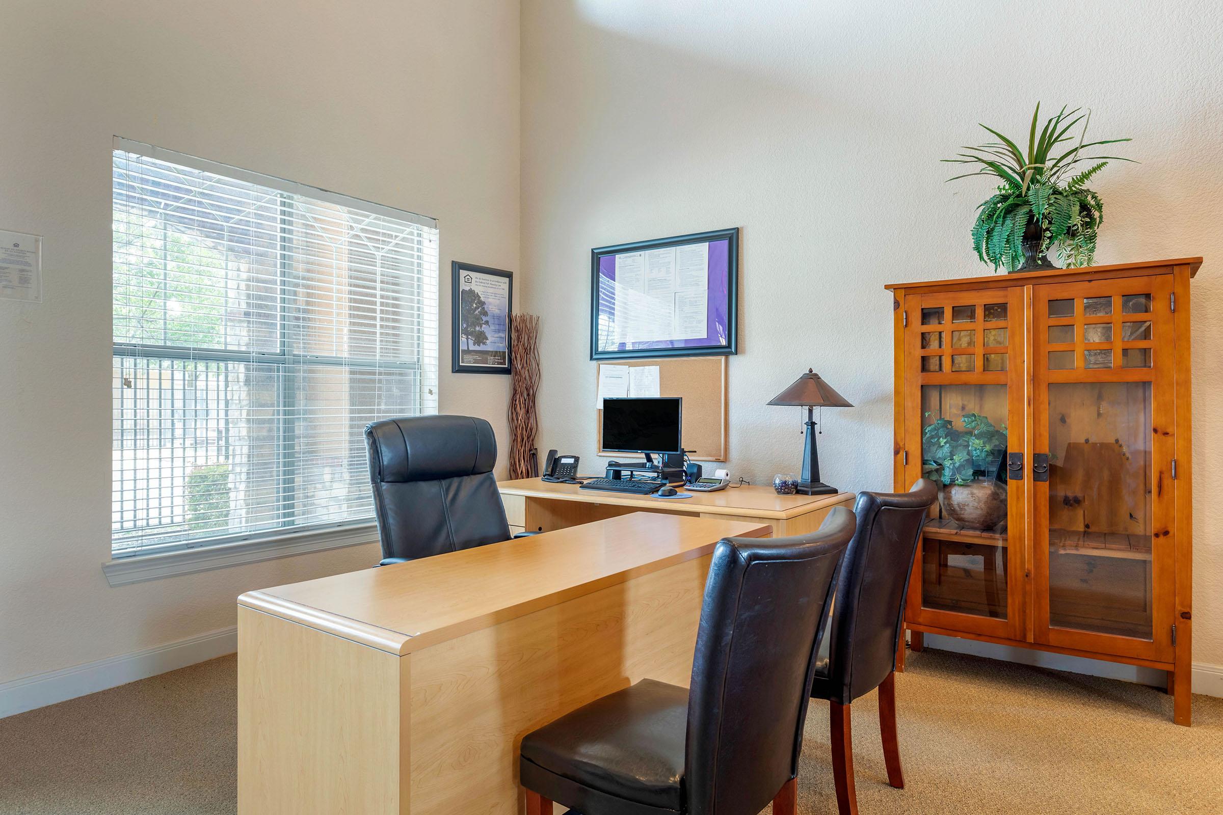A modern office space featuring a wooden desk and a black ergonomic chair. To the right, there is a glass-front cabinet displaying decorative items. A desk lamp sits on the desk, and a computer monitor is visible. Large windows let in natural light, with greenery visible outside. The walls are light-colored.