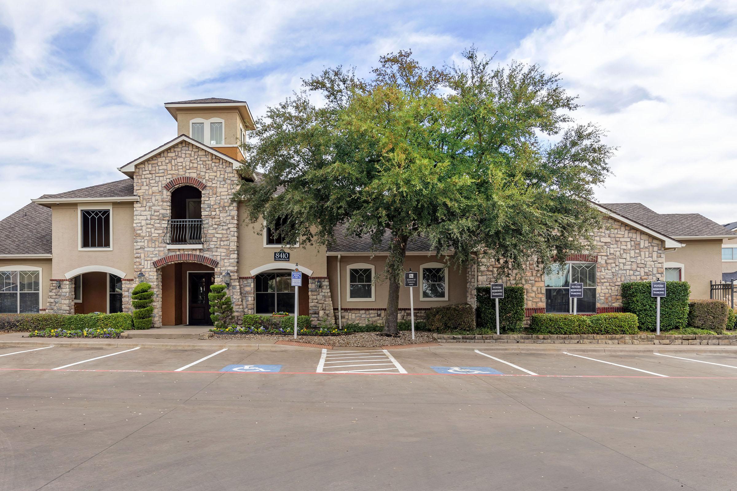 A residential building featuring a stone facade and multiple arched windows. The structure includes a tower-like section with a peaked roof. Surrounding the building are neatly trimmed hedges, flower beds, and parking spaces, including designated spots for disabled parking. The sky is partly cloudy.