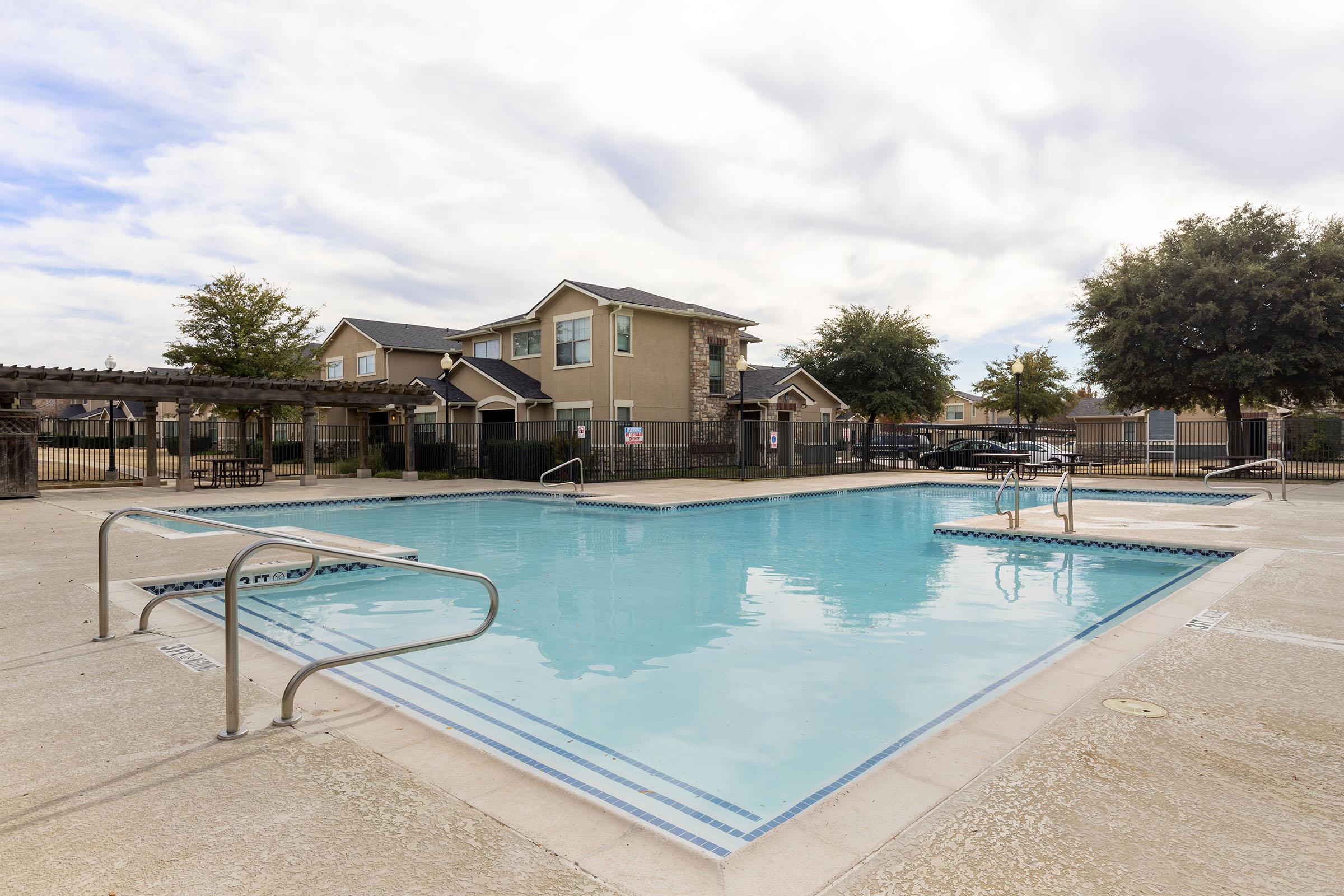 A clear swimming pool surrounded by concrete, with ladder access on one side. In the background, there are residential buildings and trees. The sky is partly cloudy, creating a tranquil atmosphere around the pool area.
