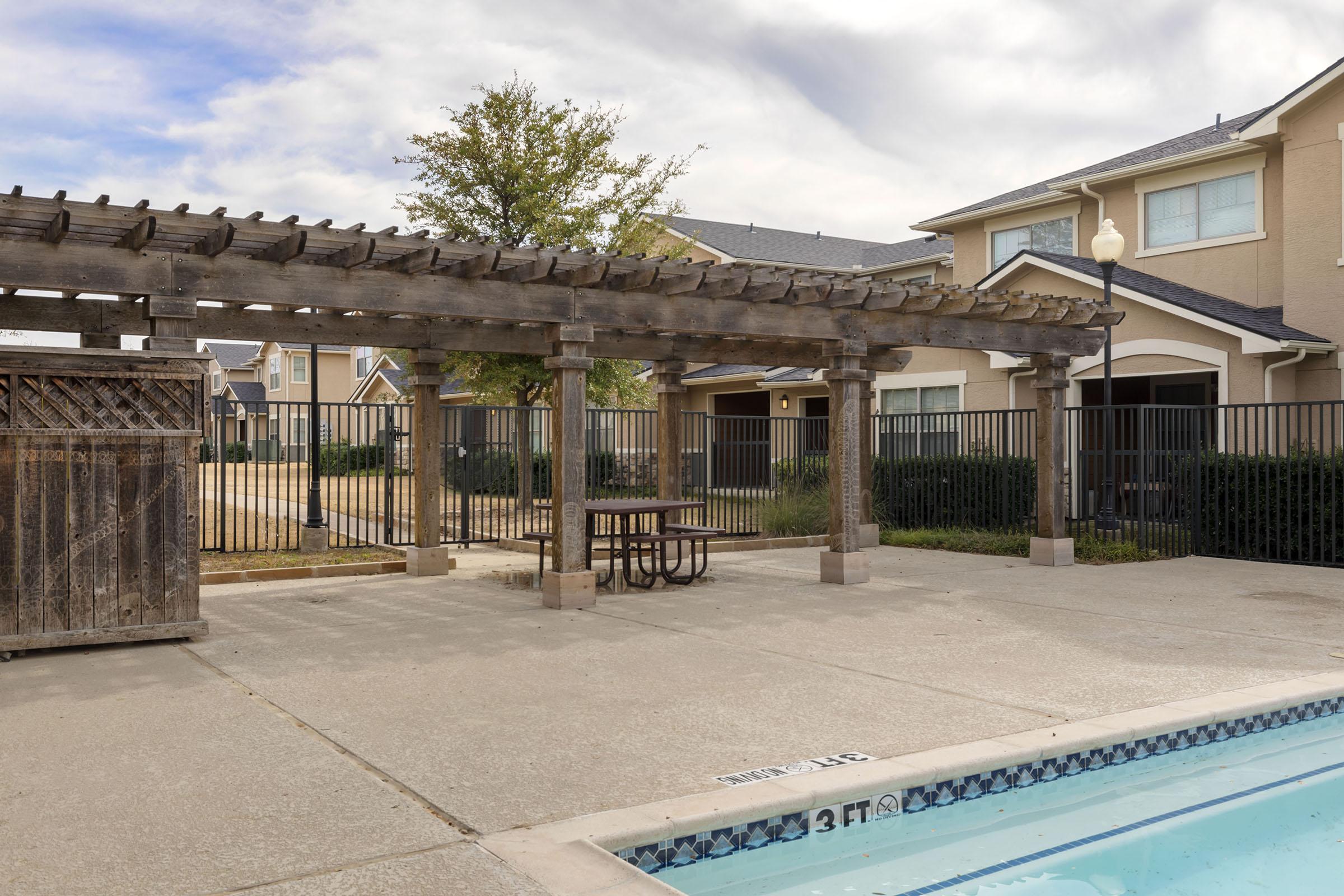 A view of a pool area featuring a pergola with wooden beams, a picnic table underneath, and a fenced-in space. In the background, there are residential buildings and landscaped greenery. The pool has markings showing the depth, with a clear sky overhead.