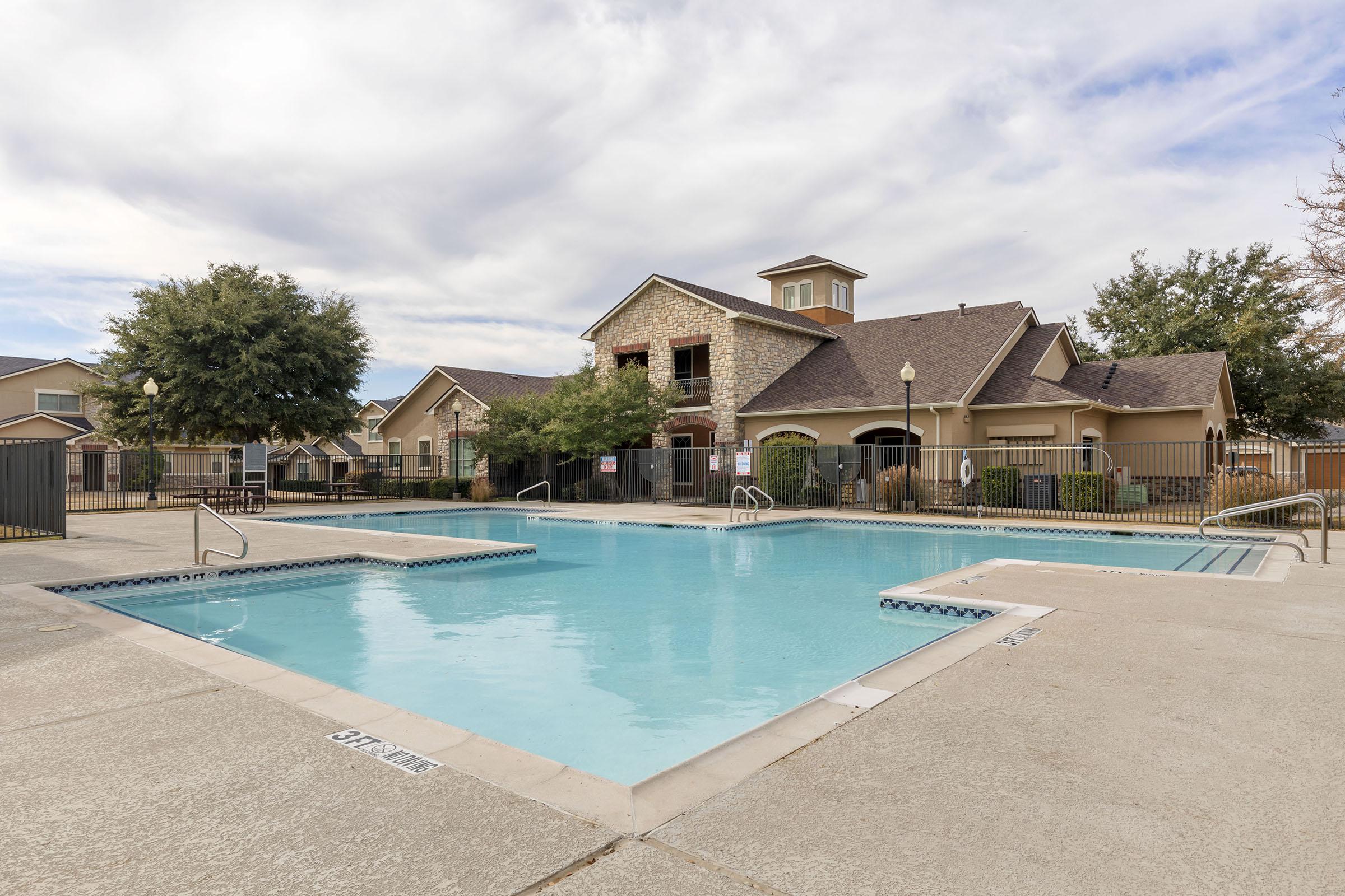 A spacious outdoor swimming pool surrounded by a fenced area, with a clubhouse visible in the background. The pool features a shallow end with stairs and is bordered by trees and well-maintained landscaping. The sky is partly cloudy, indicating a pleasant day.