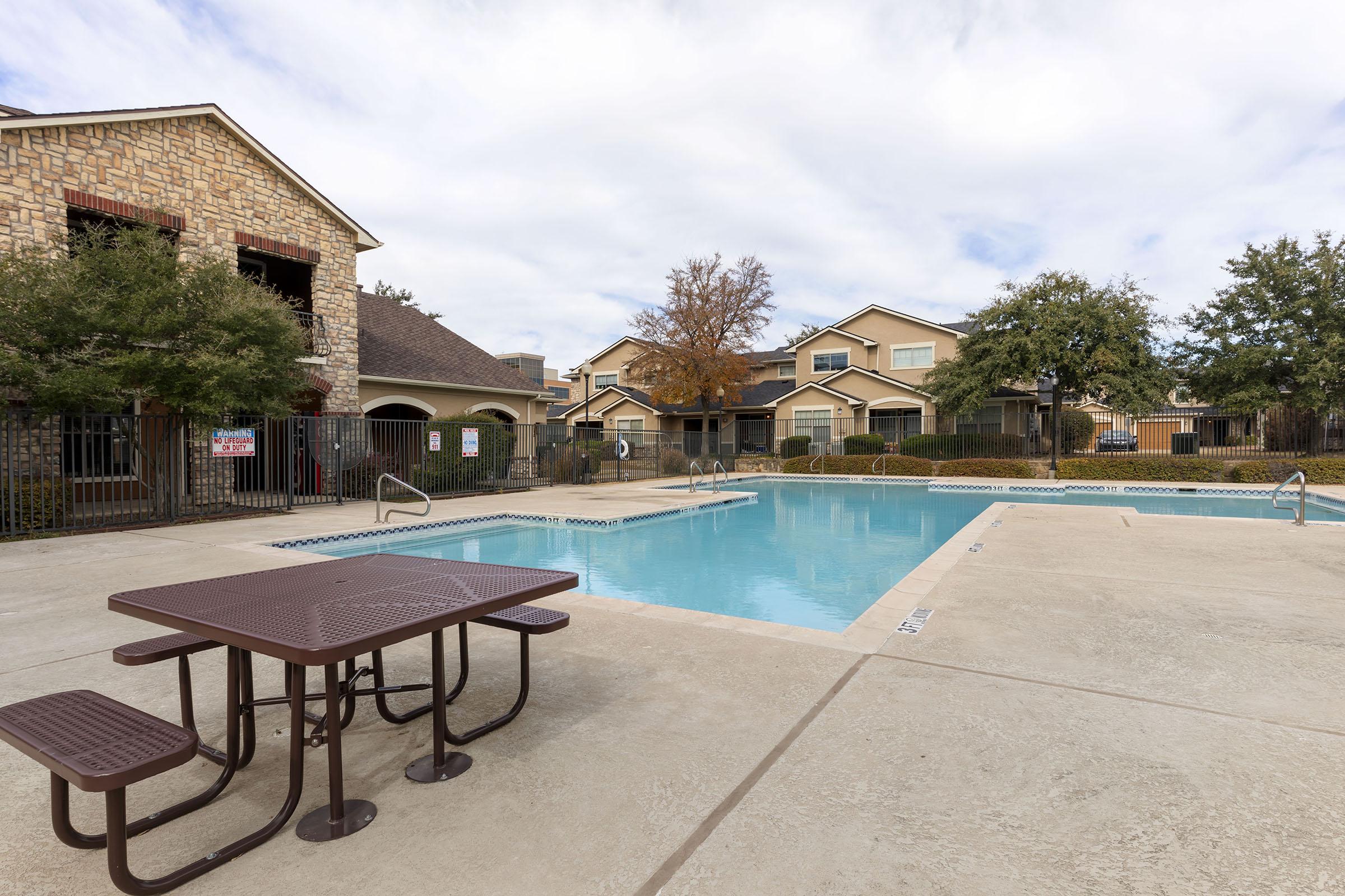 A clear swimming pool surrounded by a concrete deck, with a picnic table nearby. The pool area features lounge chairs and is enclosed by a fence. Residential buildings with balconies are visible in the background, under a partly cloudy sky.