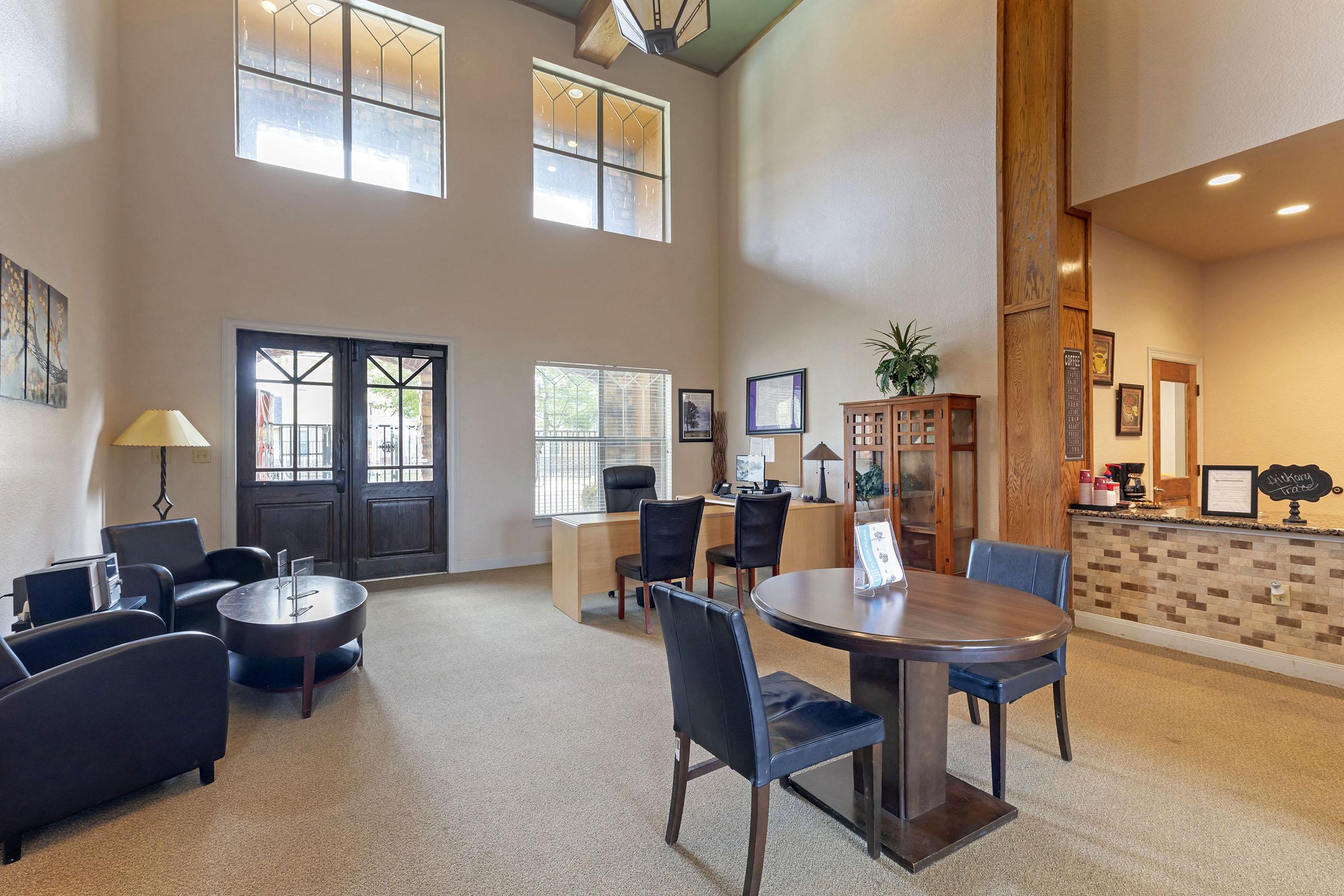 Interior view of a welcoming reception area with high ceilings. Features include a desk with a computer, a round table with chairs, dark armchairs, and a lamp. Large windows allow natural light, creating a warm atmosphere. Wall decor includes paintings, enhancing the aesthetic appeal of the space.