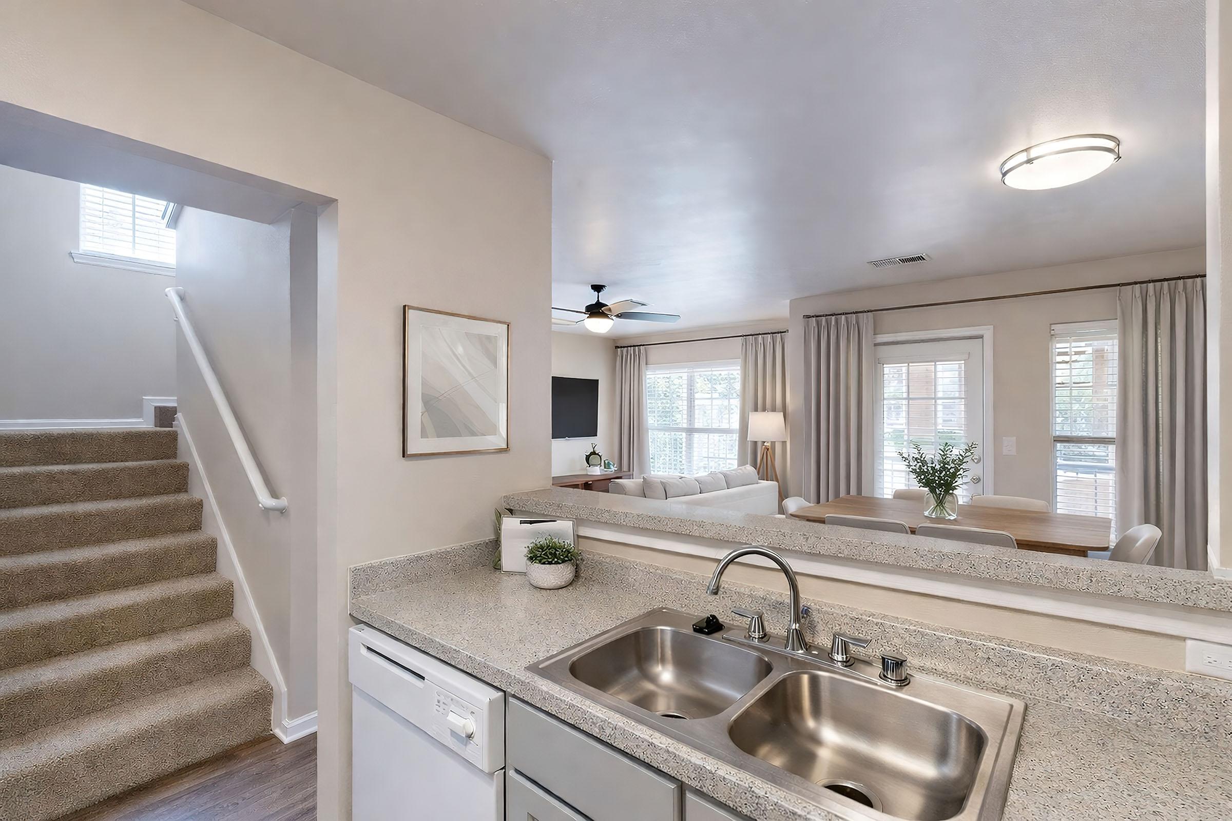 A modern kitchen view featuring a double sink with a dishwasher and a counter overlooking a cozy living room with a couch and TV. Stairs lead to the upper level, and large windows provide natural light. Neutral-colored walls and décor enhance the space's airy feel.