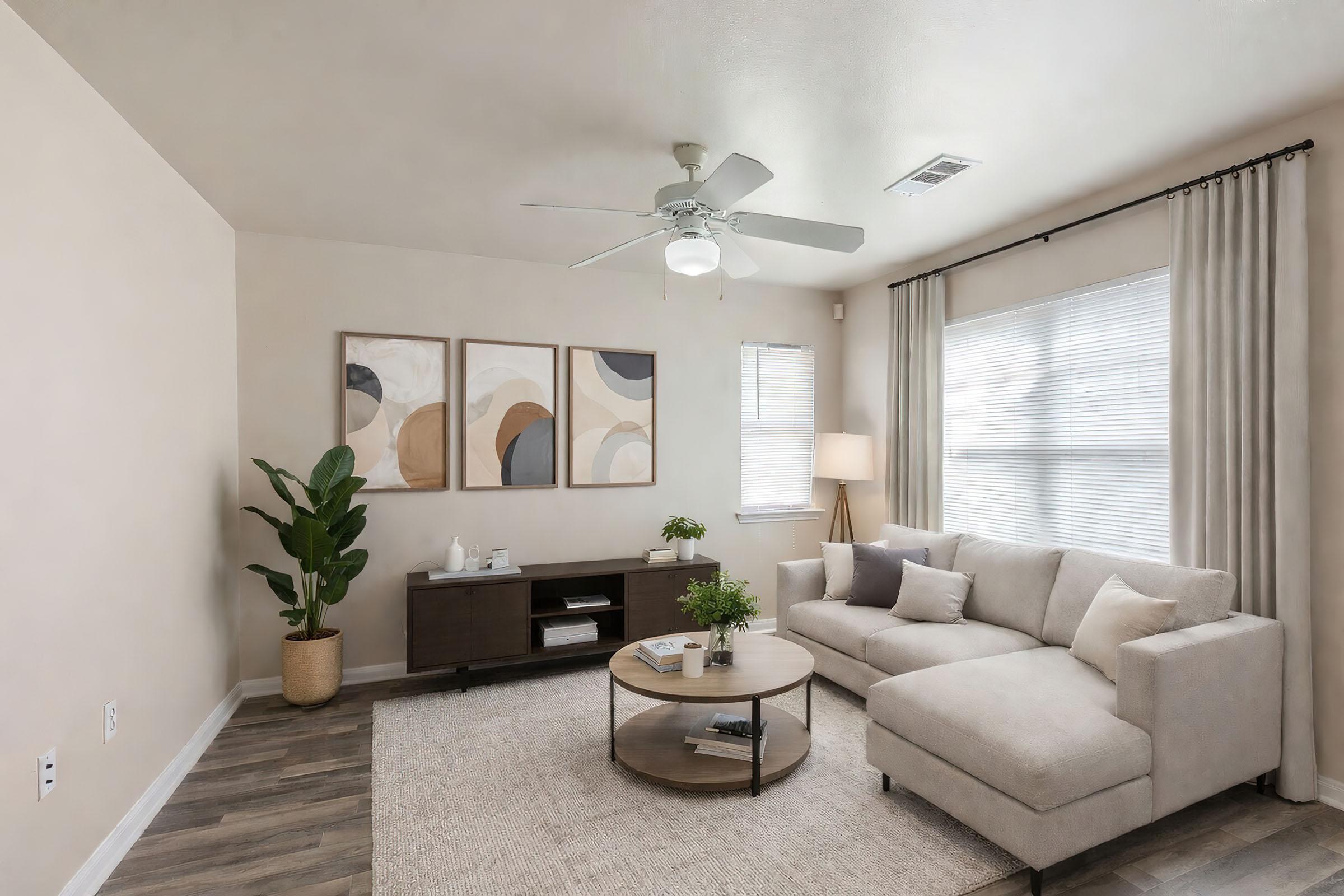 A cozy living room featuring a light beige sectional sofa, a round coffee table, and modern decor. There are three abstract framed artworks on the wall, a potted plant in the corner, and a TV stand with minimal accessories. Soft natural light filters through the window, enhancing the inviting atmosphere.