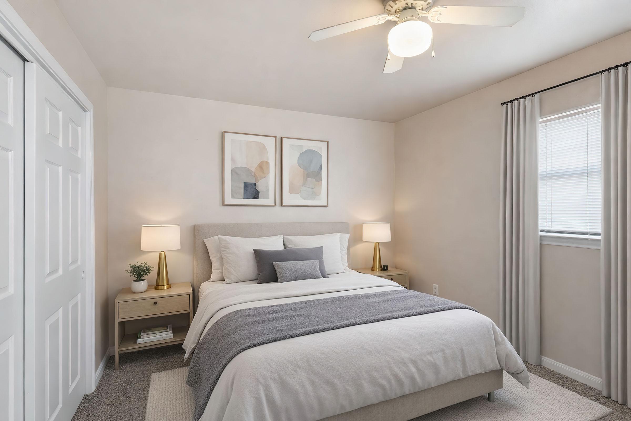 A cozy bedroom featuring a light-colored wall, a queen-sized bed with a gray blanket and decorative pillows, two bedside lamps on wooden nightstands, and abstract artwork above the bed. The room has a ceiling fan and a window with sheer curtains, creating a warm and inviting atmosphere.