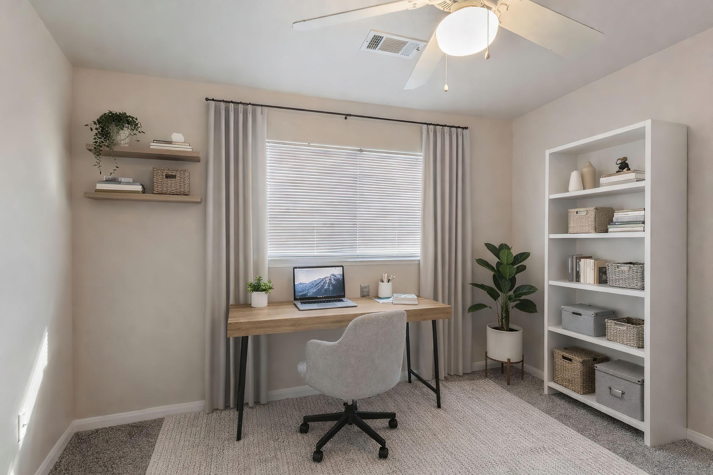 A cozy home office featuring a wooden desk with a computer, a comfortable gray chair, a large window with blinds, and light curtains. Shelves are mounted on the wall, displaying decorative items and storage baskets. A potted plant adds a touch of greenery. The room has a neutral color palette and soft carpeting.
