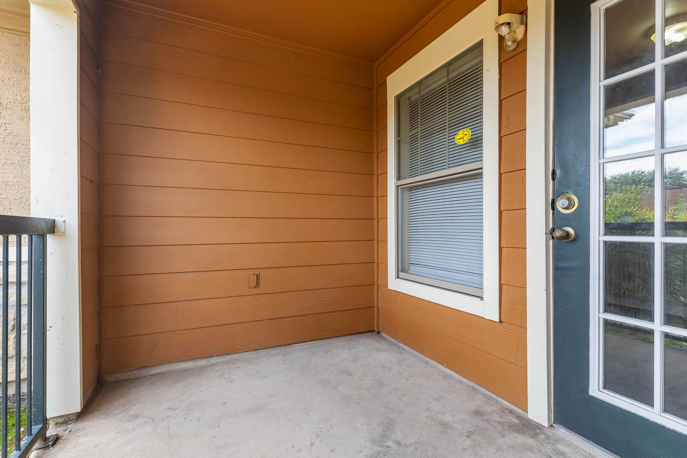A small balcony with brown wooden siding and a concrete floor. There is a door with clear glass panels on the right side and a window on the left. The balcony is empty, providing a simple and peaceful outdoor space. Natural light filters in, highlighting the warm tones of the wood.