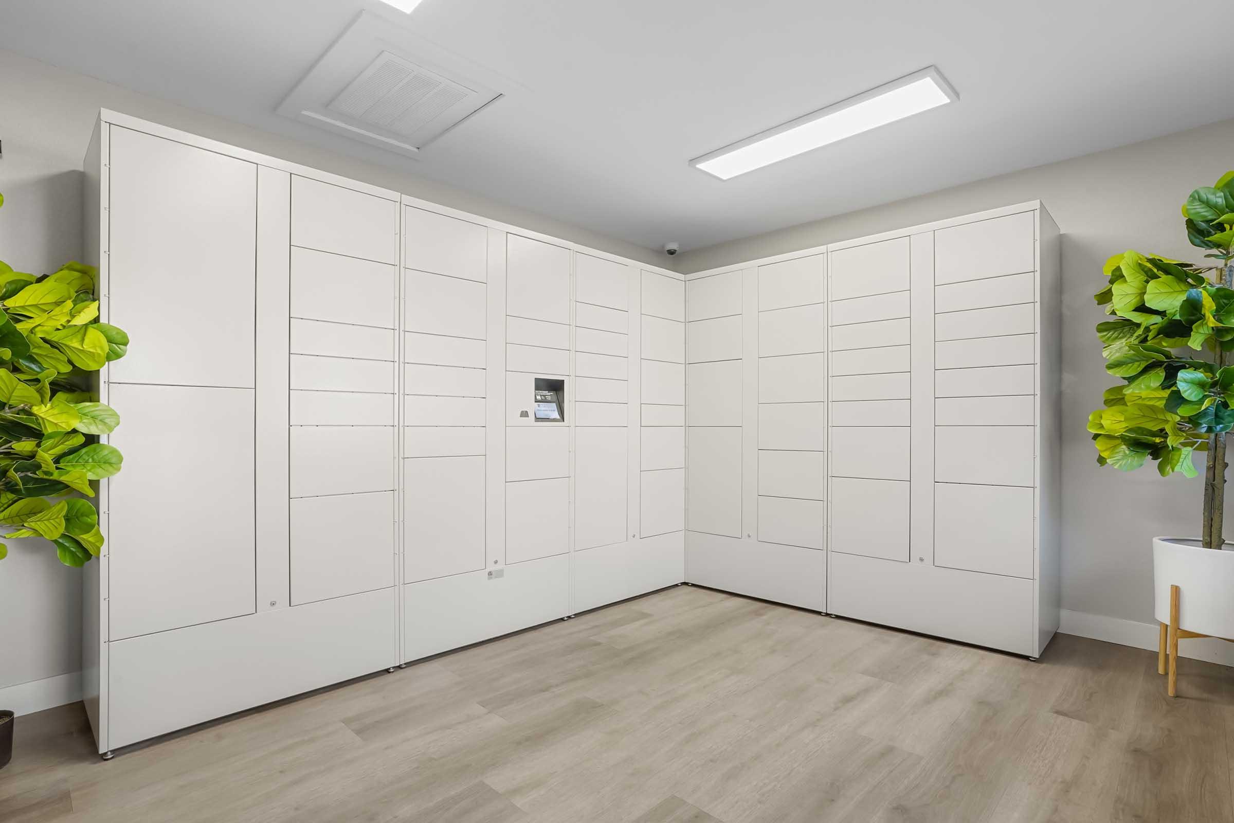 A modern room featuring a set of white package lockers against a light-colored wall. There are potted green plants in the corners, adding a touch of greenery to the space. The floor is made of light wooden panels, and overhead lighting provides bright illumination.