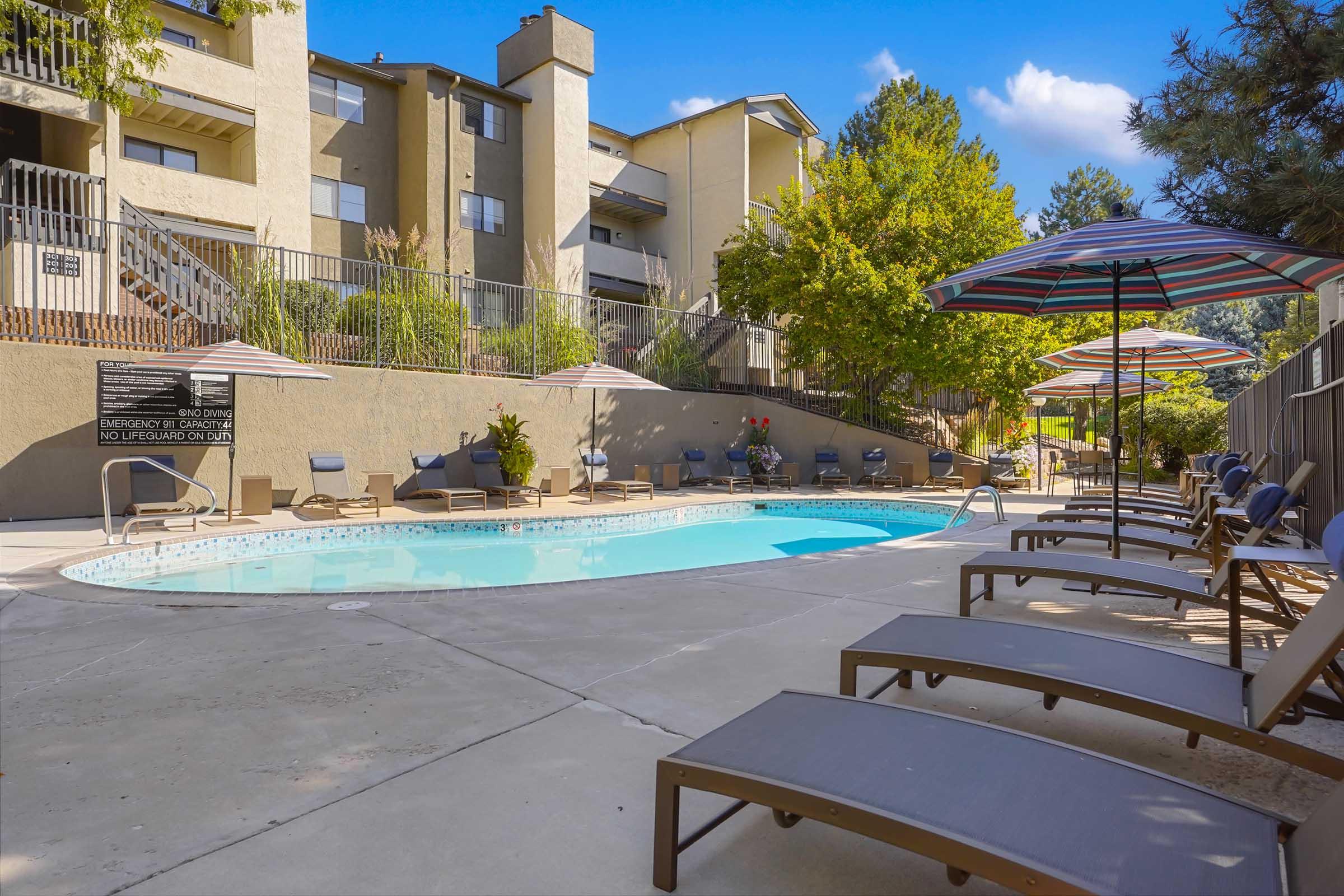 A tranquil swimming pool area surrounded by lounge chairs and umbrellas, with a tan-colored building and lush greenery in the background. The pool is inviting, reflecting the clear blue sky, while the patio features shaded seating for relaxation.