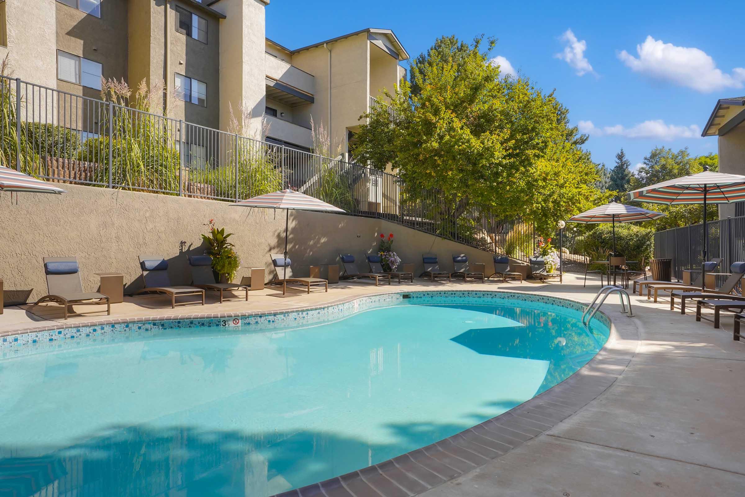 A clear swimming pool surrounded by lounge chairs and umbrellas, set against a backdrop of apartment buildings and lush greenery under a bright blue sky. The area features well-maintained landscaping, creating a relaxing outdoor space.