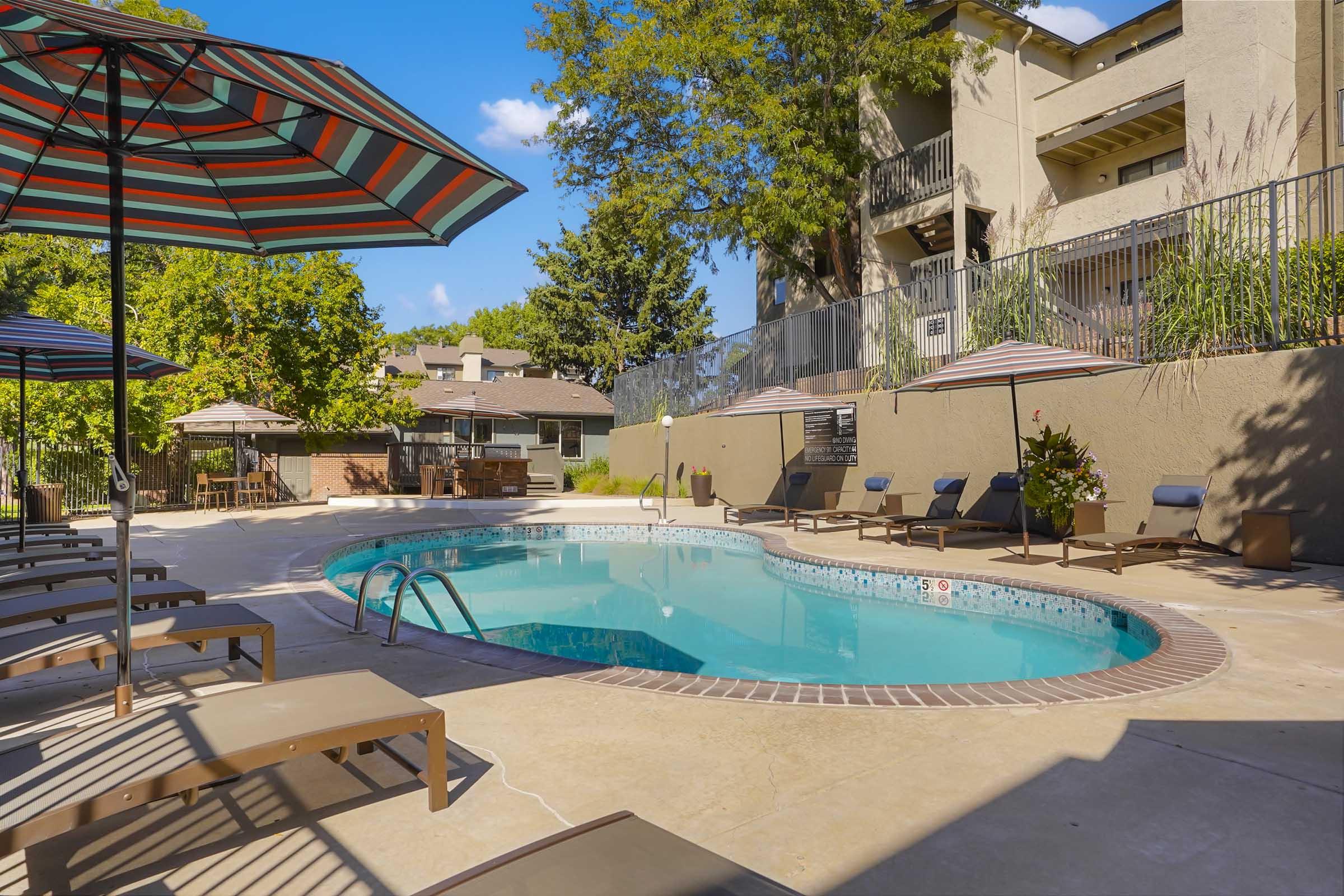A serene pool area featuring a clear blue swimming pool surrounded by sun loungers and large umbrellas. Green trees and a building are visible in the background under a bright blue sky, creating a relaxing atmosphere for leisure and enjoyment.