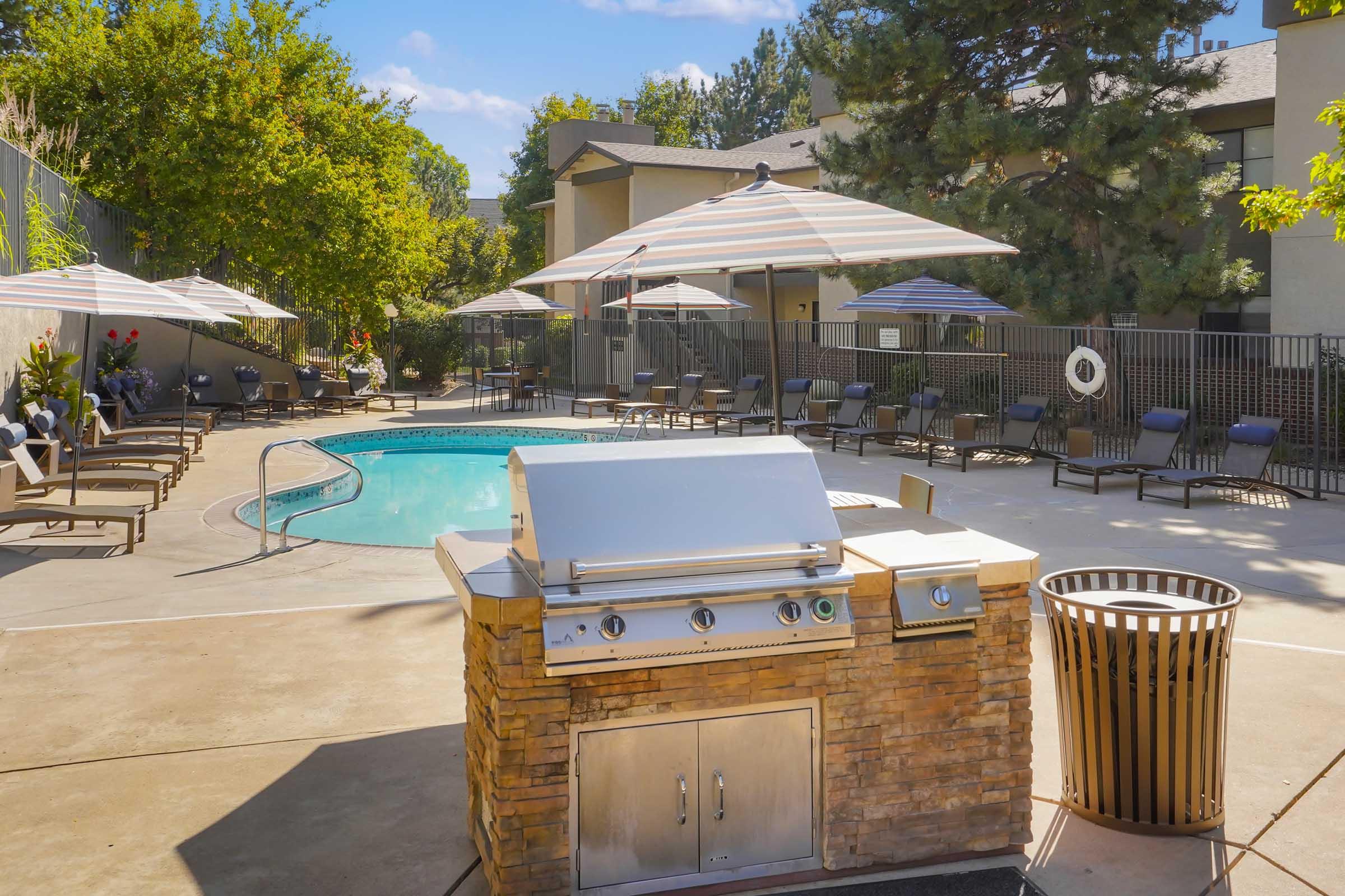 A modern outdoor pool area featuring a grill station made of stone, lounge chairs, and umbrellas. The pool is surrounded by greenery and apartment buildings in the background, creating a relaxing atmosphere for residents.