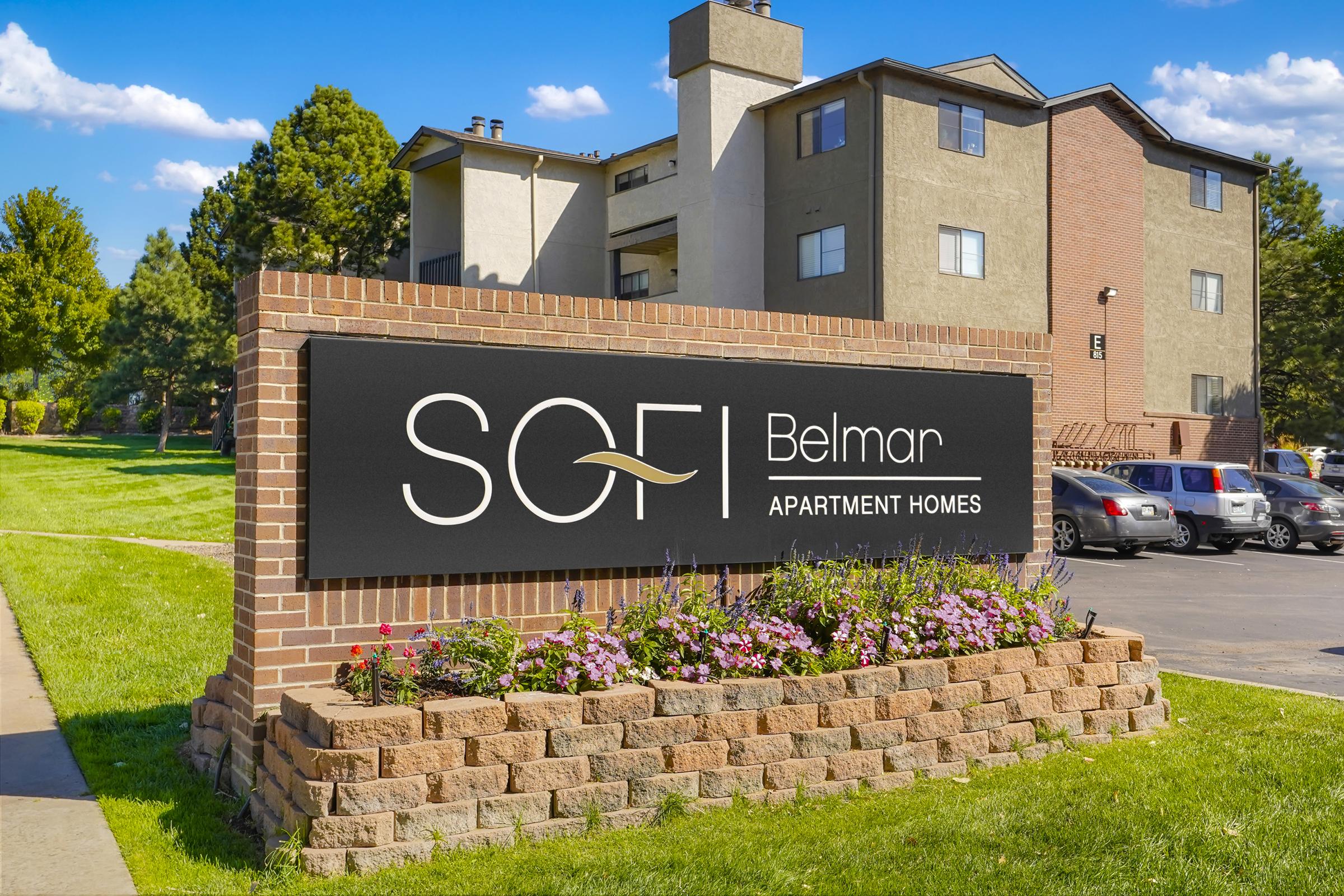 Sign for SOFI Belmar Apartment Homes, featuring a modern design with the name prominently displayed. The background shows well-maintained apartment buildings and greenery, with flowers planted at the base of the sign and cars parked nearby. The sky is clear with a few clouds.