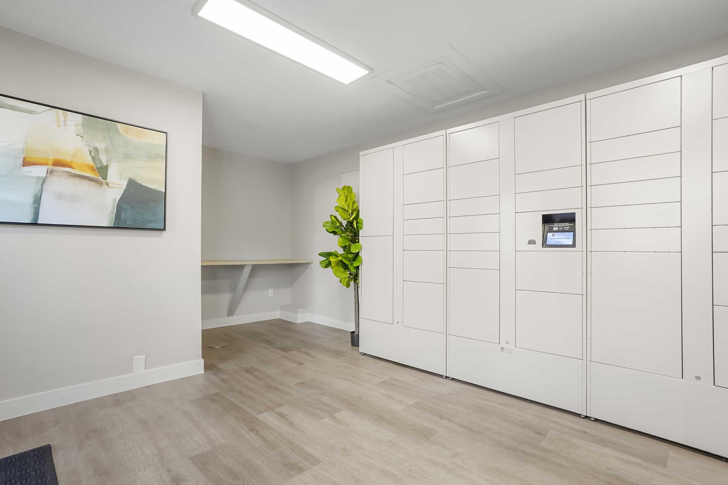 Modern interior space featuring a wall-mounted art piece, a large package locker system, and a potted plant. The room has light-colored walls and wooden flooring, with a long countertop along one side, creating a clean and organized environment. Natural light enters, enhancing the inviting atmosphere.