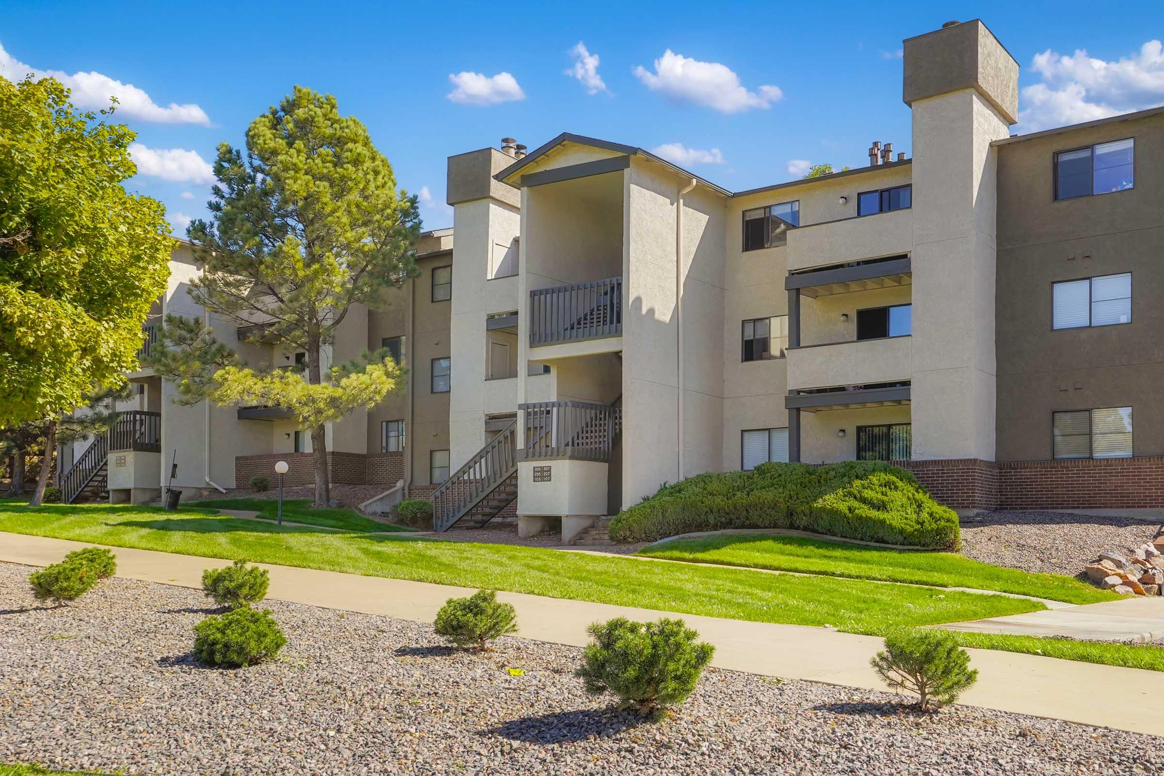 A modern apartment building with three stories, featuring a central balcony and multiple windows. Surrounding the building are well-maintained lawns and small shrubs along a pathway, under a clear blue sky with fluffy white clouds. The scene conveys a clean and inviting residential atmosphere.