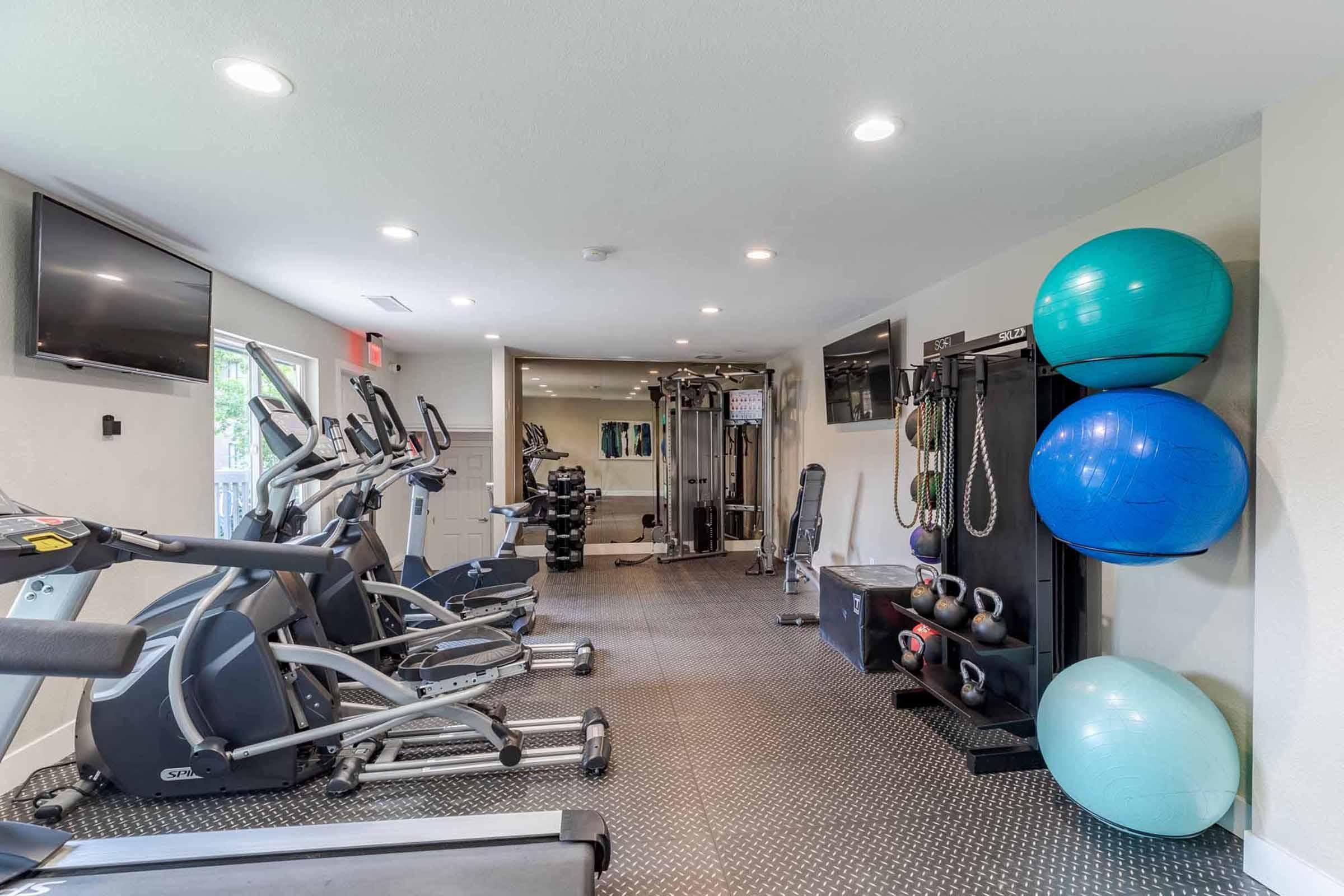 A well-lit gym space featuring various fitness equipment such as elliptical machines, weights, and exercise balls. The room has a mirror and multiple large windows for natural light, creating an inviting atmosphere for workouts.
