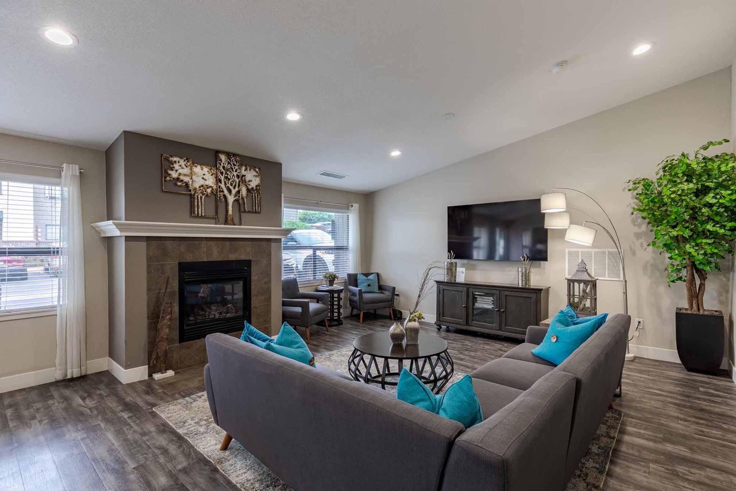 A modern living room featuring a gray sofa with turquoise cushions, a round coffee table, and a decorative fireplace. A large TV is mounted on the wall, and there's a potted plant in a corner. Natural light comes through windows, and artwork is displayed above the fireplace, creating a warm and inviting atmosphere.