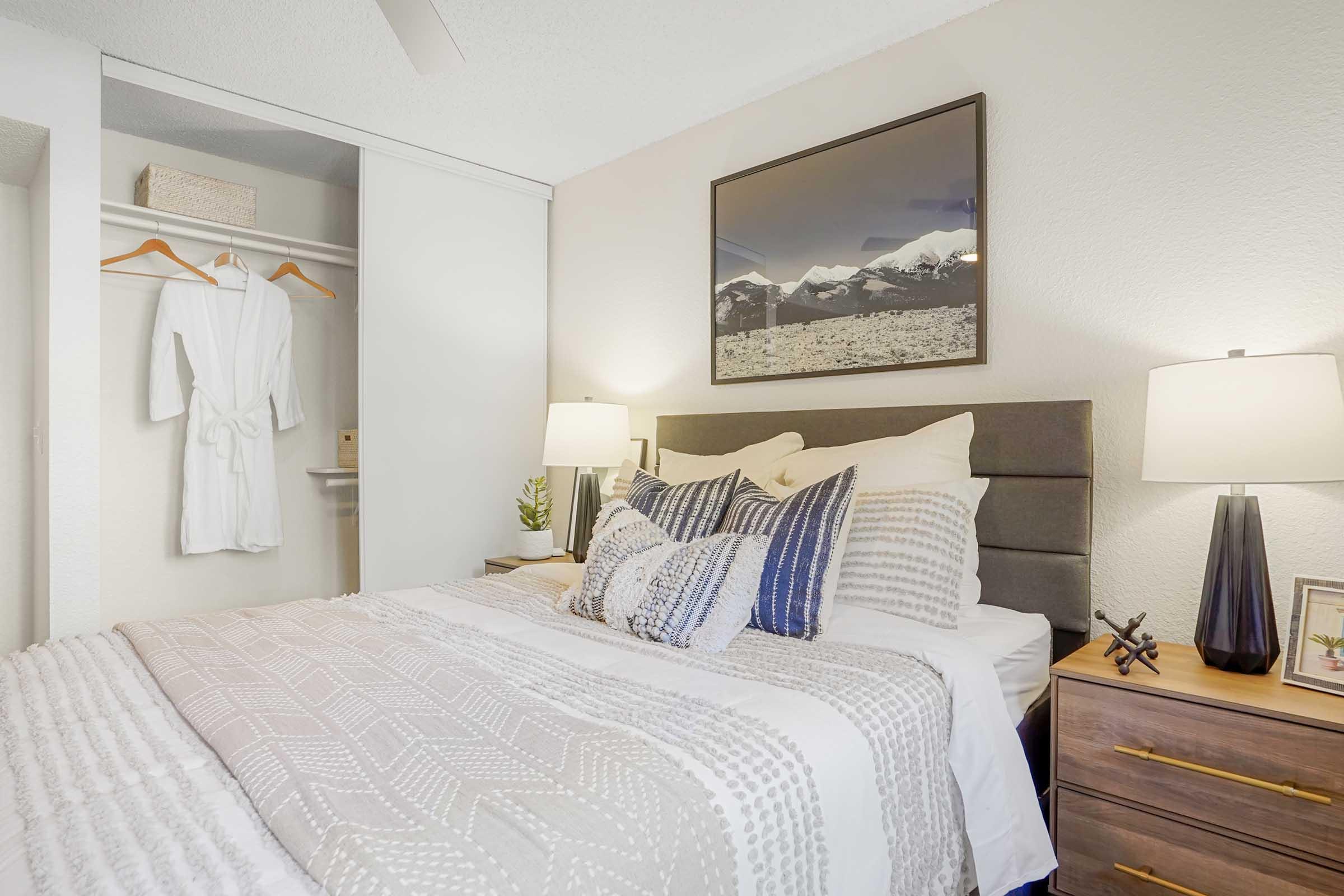 Cozy bedroom featuring a neatly made bed with decorative pillows, bedside tables with lamps, a mountain-themed artwork, and a closet with a white robe hanging. Soft lighting creates a warm atmosphere, and a small plant adds a touch of greenery to the space.