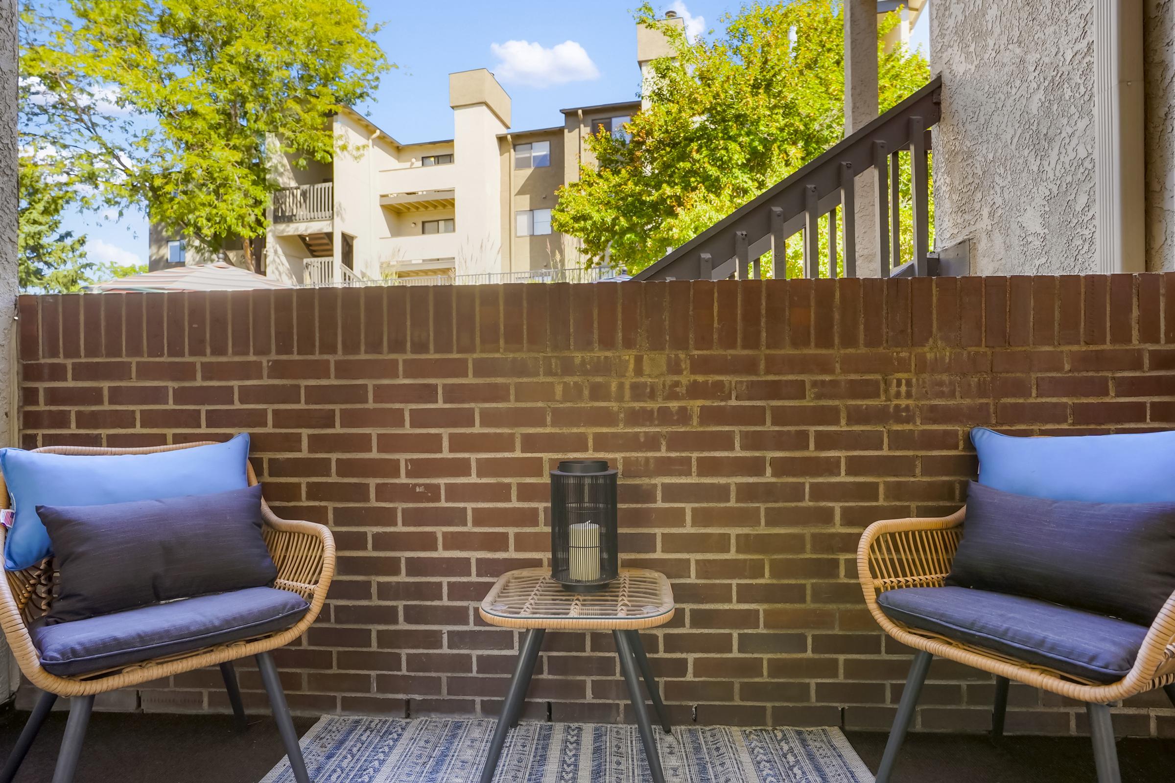 Cozy outdoor balcony featuring two wicker chairs with blue cushions, a small round table in the center, and a lantern. A brick wall provides privacy, and greenery from nearby trees and apartment buildings can be seen in the background under a clear blue sky.