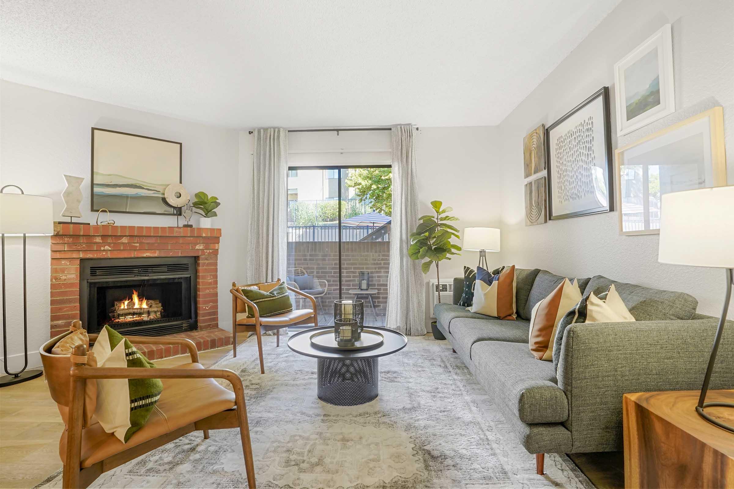 Cozy living room featuring a brick fireplace, a comfortable gray sofa with colorful pillows, two stylish chairs, and a circular coffee table. Natural light flows through large windows, and decorative art is displayed on the walls, creating a warm and inviting atmosphere.