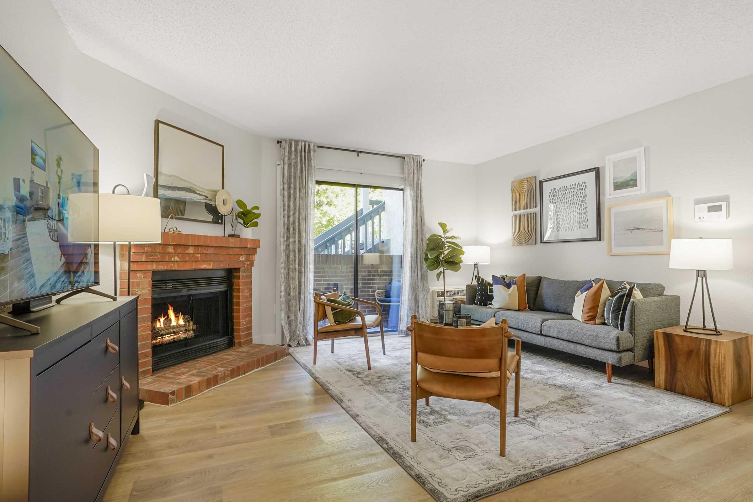 Cozy living room featuring a fireplace, a TV on a media console, and a comfortable sofa with decorative pillows. A wooden chair adds to the seating area, complemented by a rug and various wall art. Large windows let in natural light, and greenery is present, enhancing the inviting atmosphere.