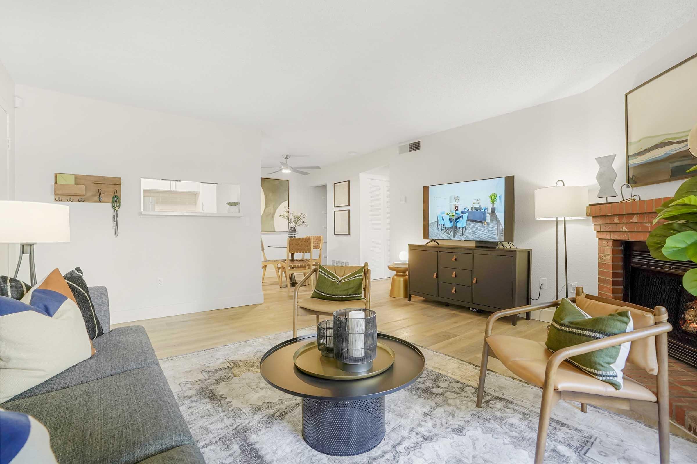 A cozy living room featuring a gray sofa with colorful cushions, a round coffee table, and a sleek TV unit. In the background, there is a dining area with a wooden table and chairs. The space is bright and airy, with a light color palette and decorative plants. A fireplace adds warmth to the setting.