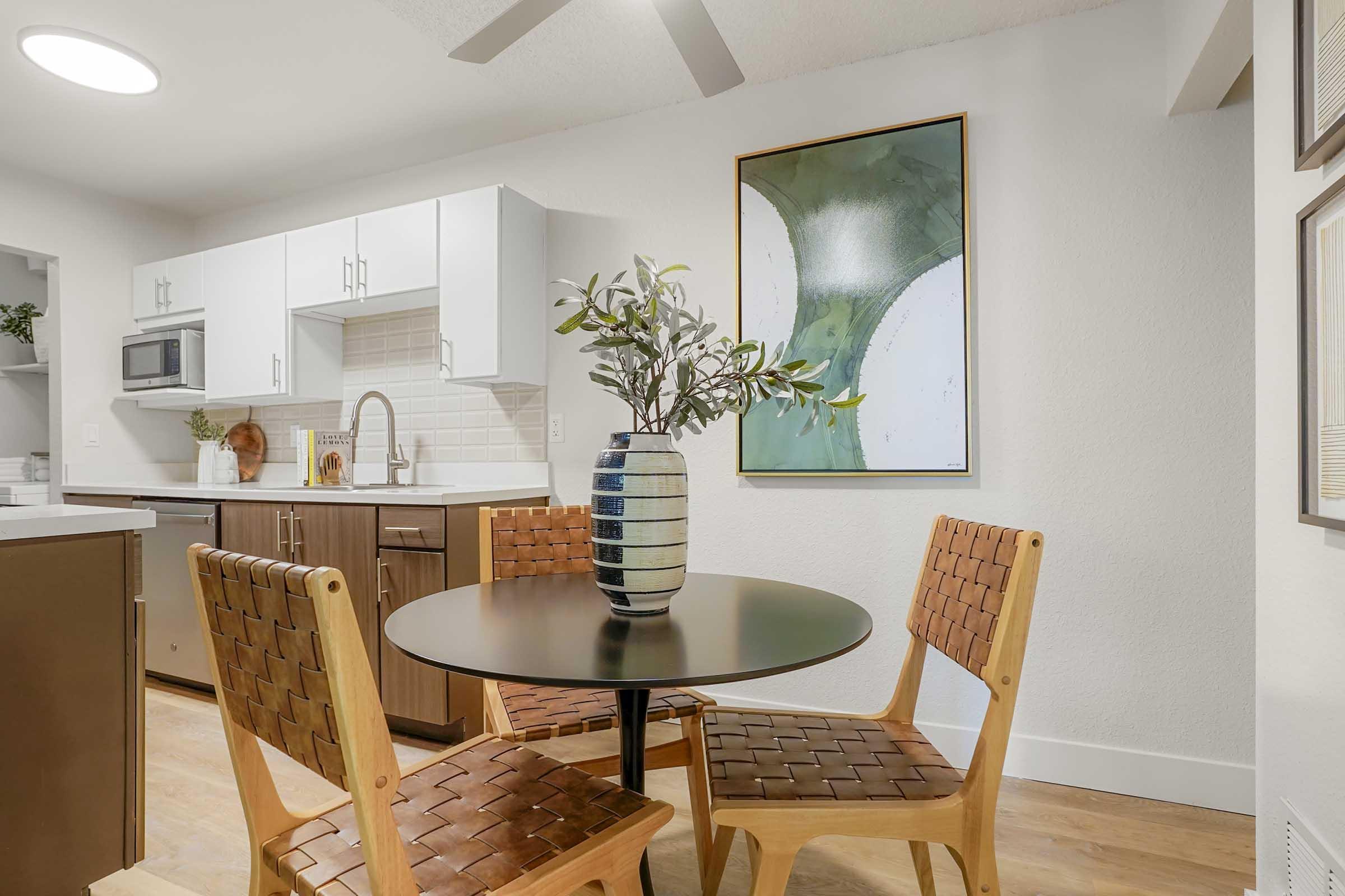 A modern kitchen area featuring a round table with woven wooden chairs. A decorative vase with greenery is on the table. The kitchen has white cabinets, a stainless steel microwave, and a minimalist artwork on the wall. Warm wooden flooring complements the space.