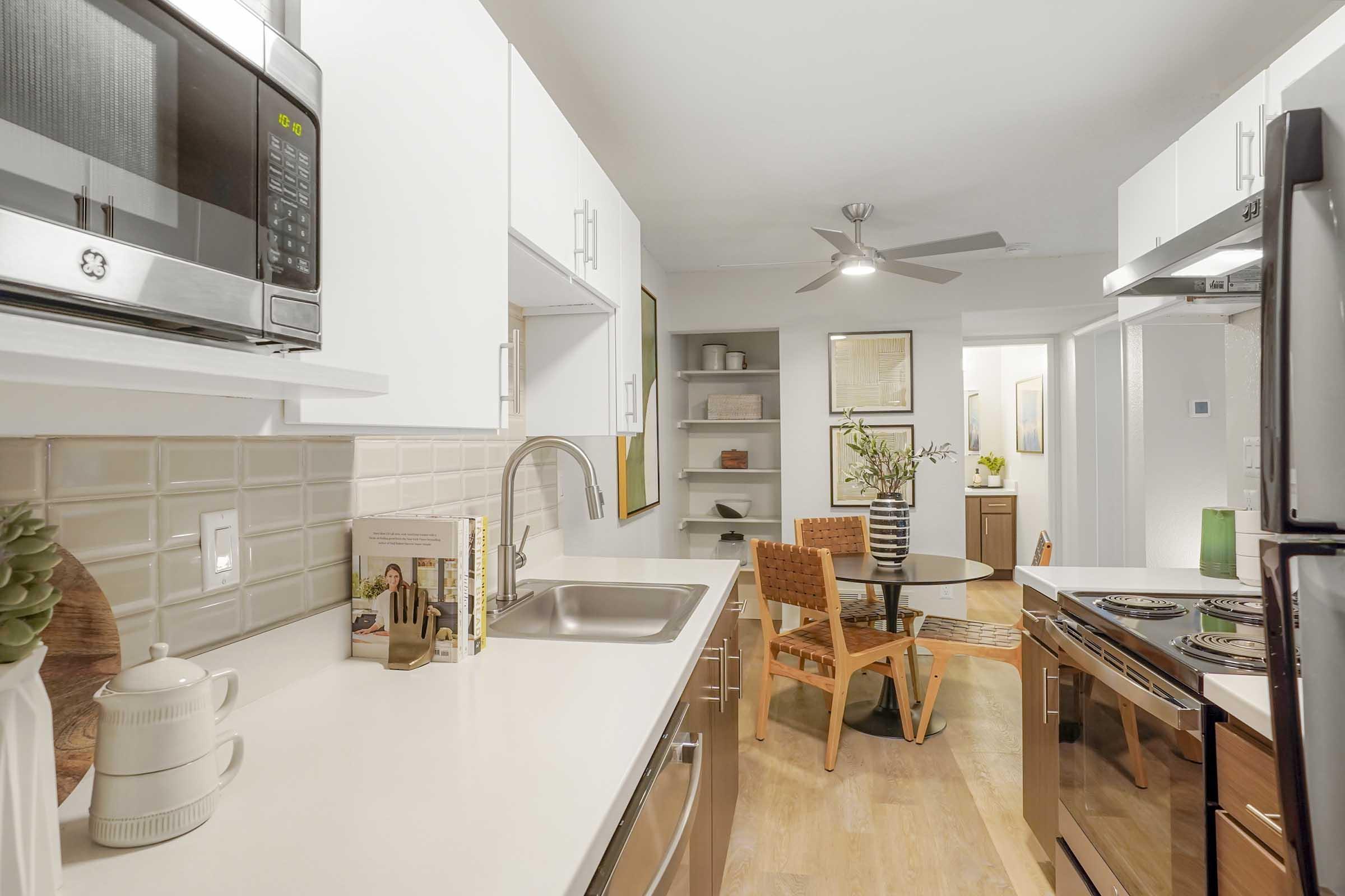 Modern kitchen featuring white cabinets, stainless steel appliances, and a sink. An open layout connects to a dining area with a round table and four chairs. Decorative elements include books and plants, with warm wooden accents and a ceiling fan providing a bright and inviting atmosphere.