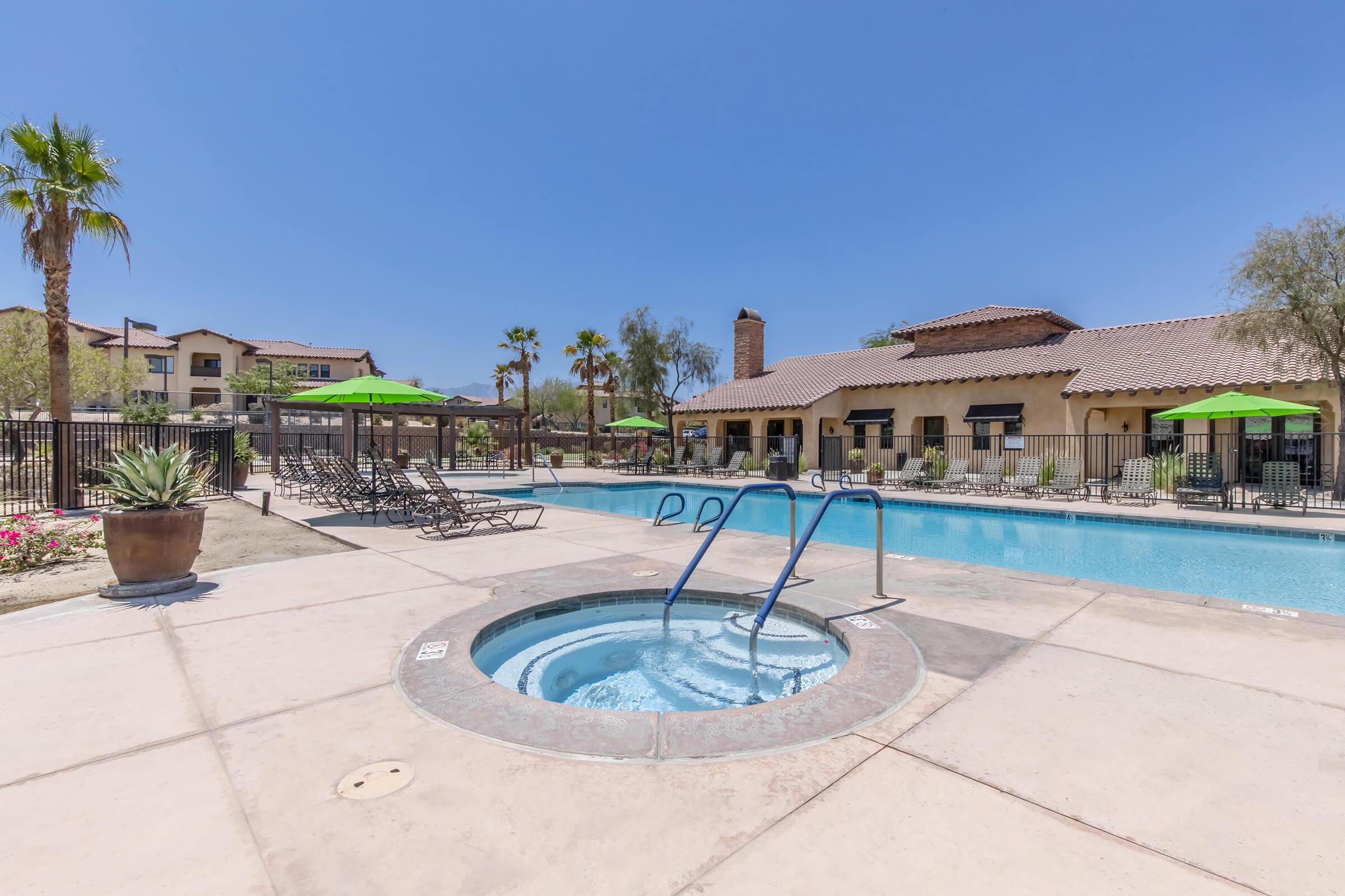 A pool area featuring a hot tub and a swimming pool surrounded by lounge chairs. Green umbrellas provide shade, and palm trees are visible in the background. A building with a tile roof is located nearby, under a clear blue sky.
