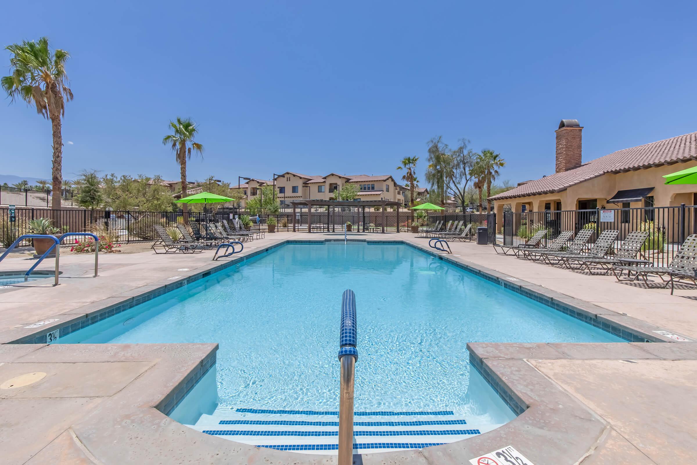 A clear blue swimming pool surrounded by sun loungers and green umbrellas, set against a backdrop of palm trees and residential buildings under a clear blue sky. The pool area is well-maintained, inviting for relaxation and leisure activities.
