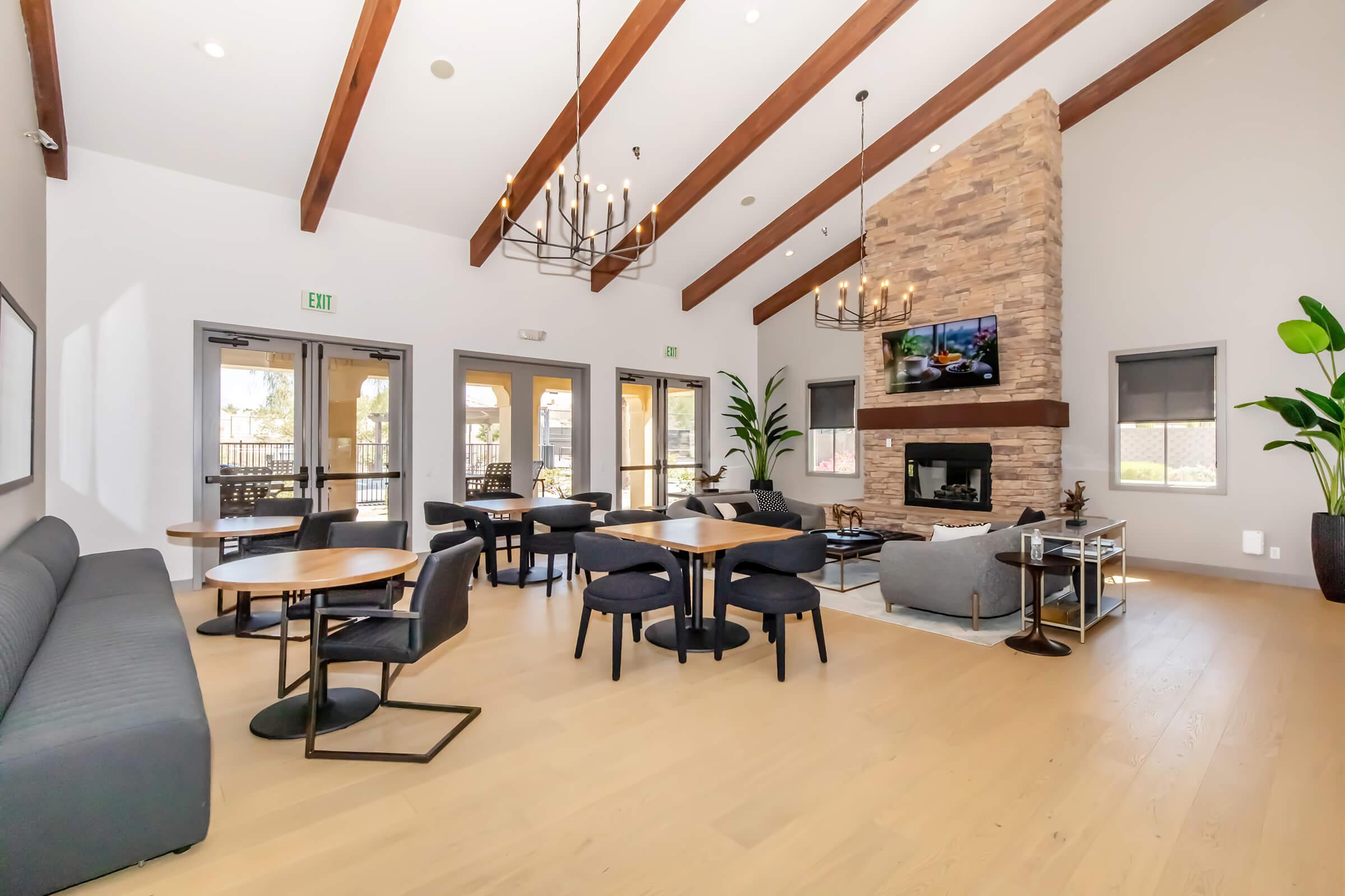 A modern living space with high wooden beams, stone fireplace, and large windows. There are several tables and chairs arranged for seating, along with a comfortable gray sofa and a television mounted on the wall. Decorative plants add a touch of greenery to the contemporary atmosphere.