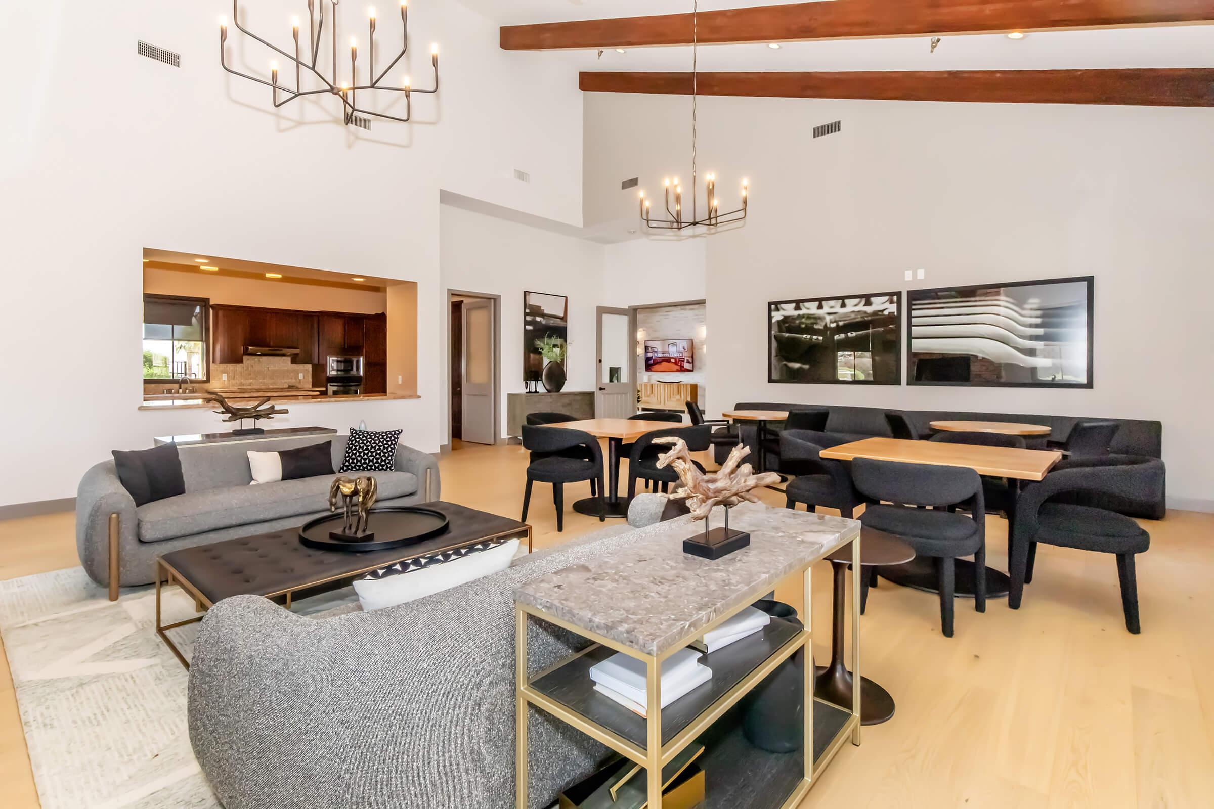 A bright, modern living space featuring a high ceiling with exposed beams. The room includes a cozy seating area with a gray sofa, a stylish coffee table, and a dining area with black chairs and wood tables. Large windows allow natural light to fill the space, creating an inviting atmosphere.