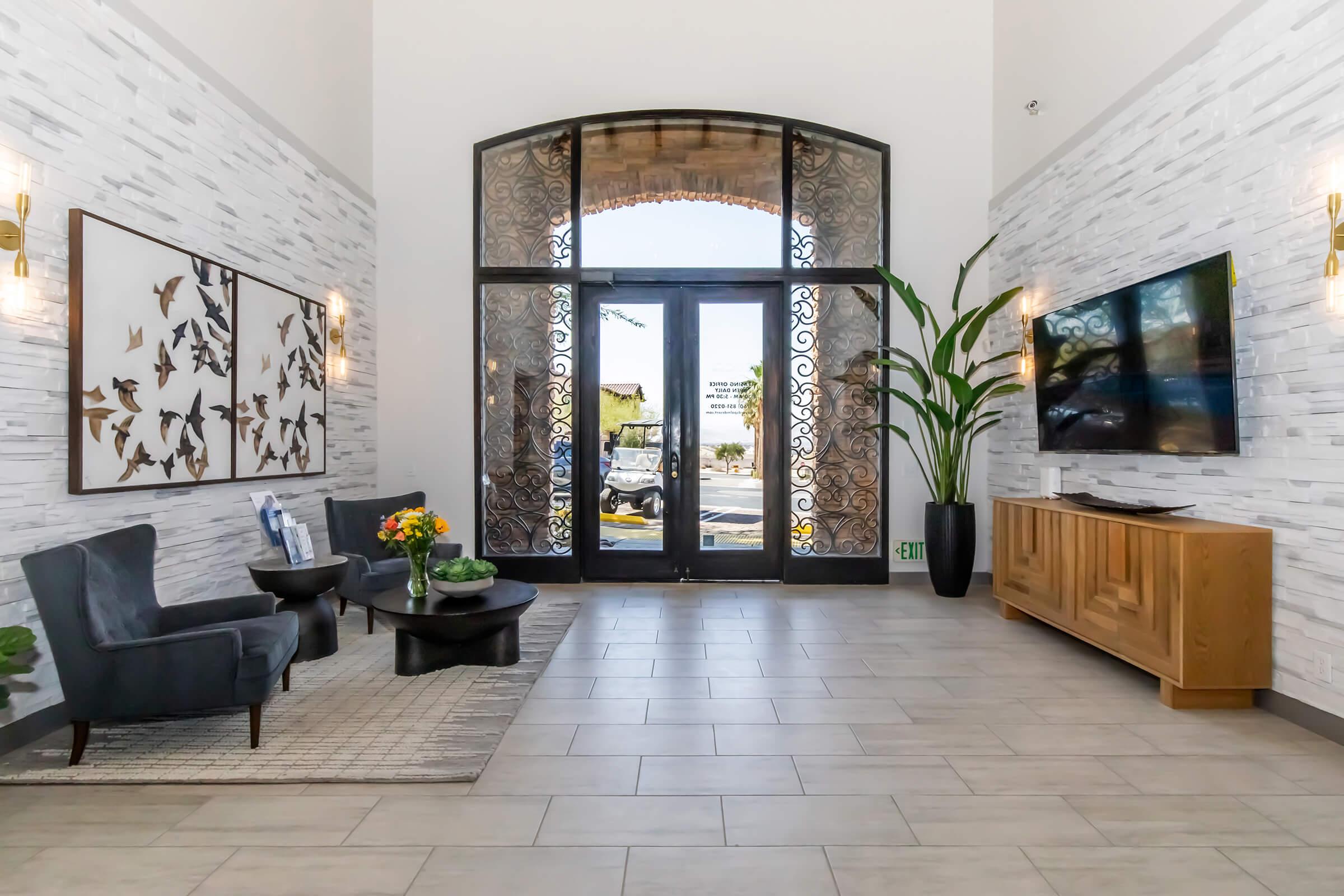 A modern lobby with a high ceiling, featuring two dark chairs and a coffee table with a flower arrangement. The walls are adorned with decorative panels and a large artwork of butterflies. Natural light streams through large glass doors, and there's a TV mounted on a wooden cabinet with plants beside it.