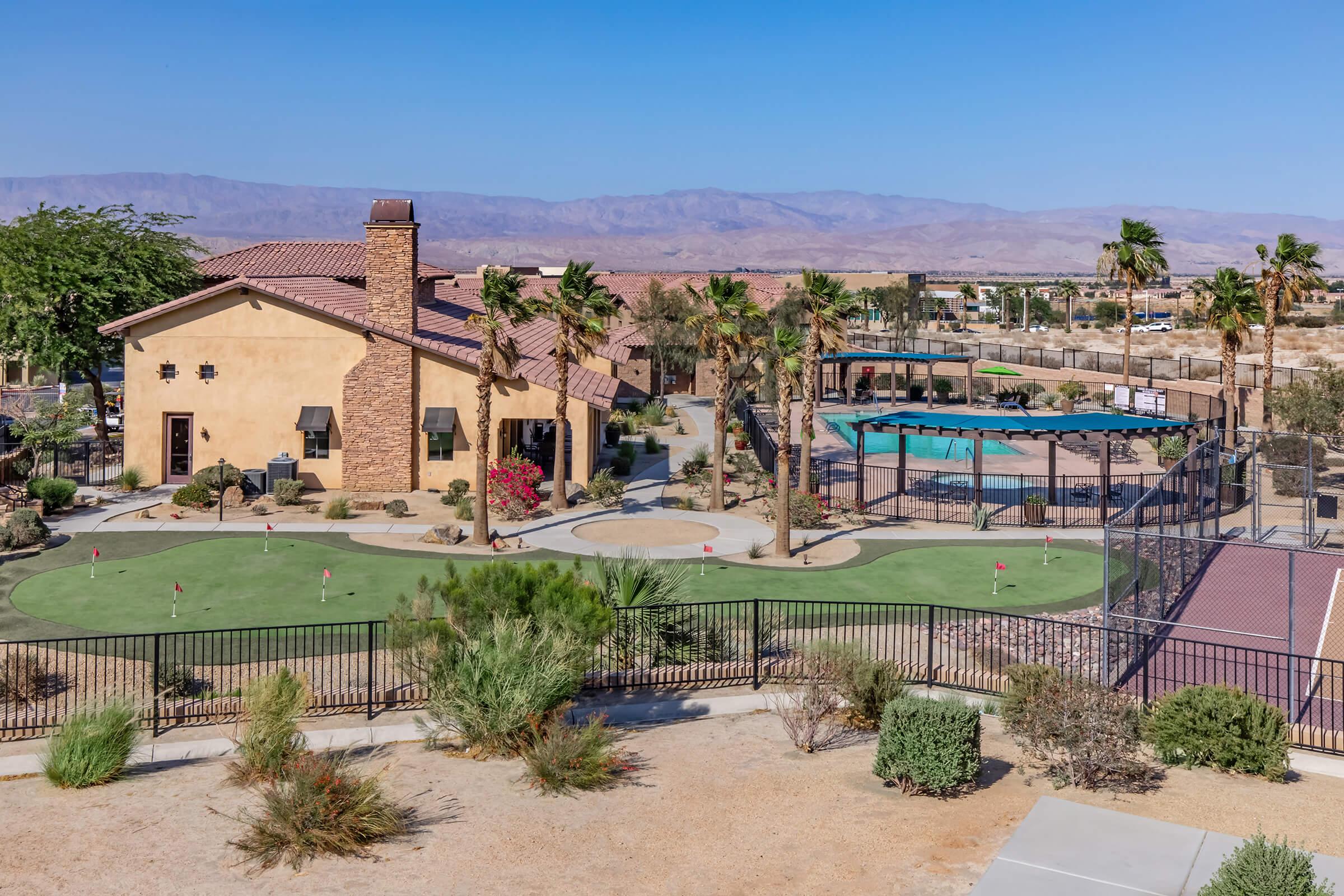 A vibrant view of a residential community featuring a modern clubhouse, a swimming pool, well-maintained landscaping with palm trees, and a golf putting green. In the background, a mountain range and blue sky complete the picturesque scene of relaxation and leisure.