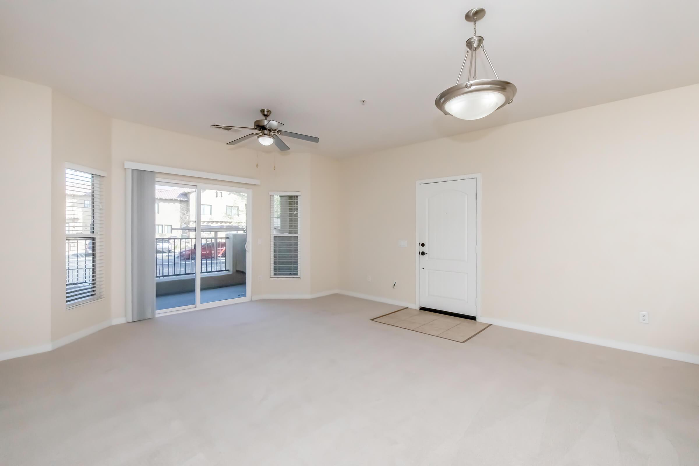 Spacious living room with beige walls and carpet, featuring a ceiling fan and light fixture. There are large windows offering natural light, and a sliding glass door leading to a balcony. The entrance door is visible, and the area is clean and empty, creating a bright and inviting space.