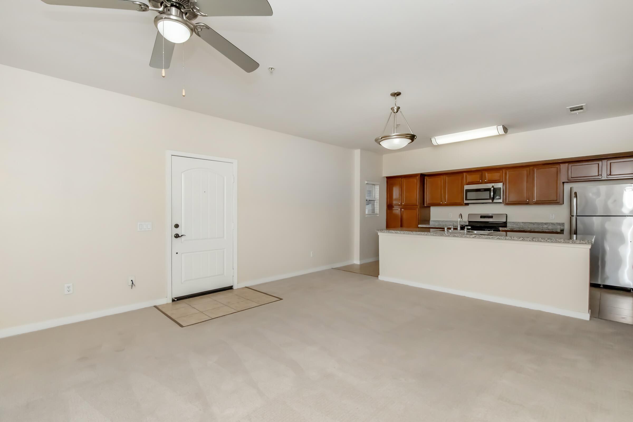 Open concept living area featuring a beige carpet, ceiling fan, and natural light from a front door. The kitchen is visible in the background with wooden cabinets, stainless steel appliances, and granite countertops. The space is neutral and inviting, ideal for a cozy living environment.