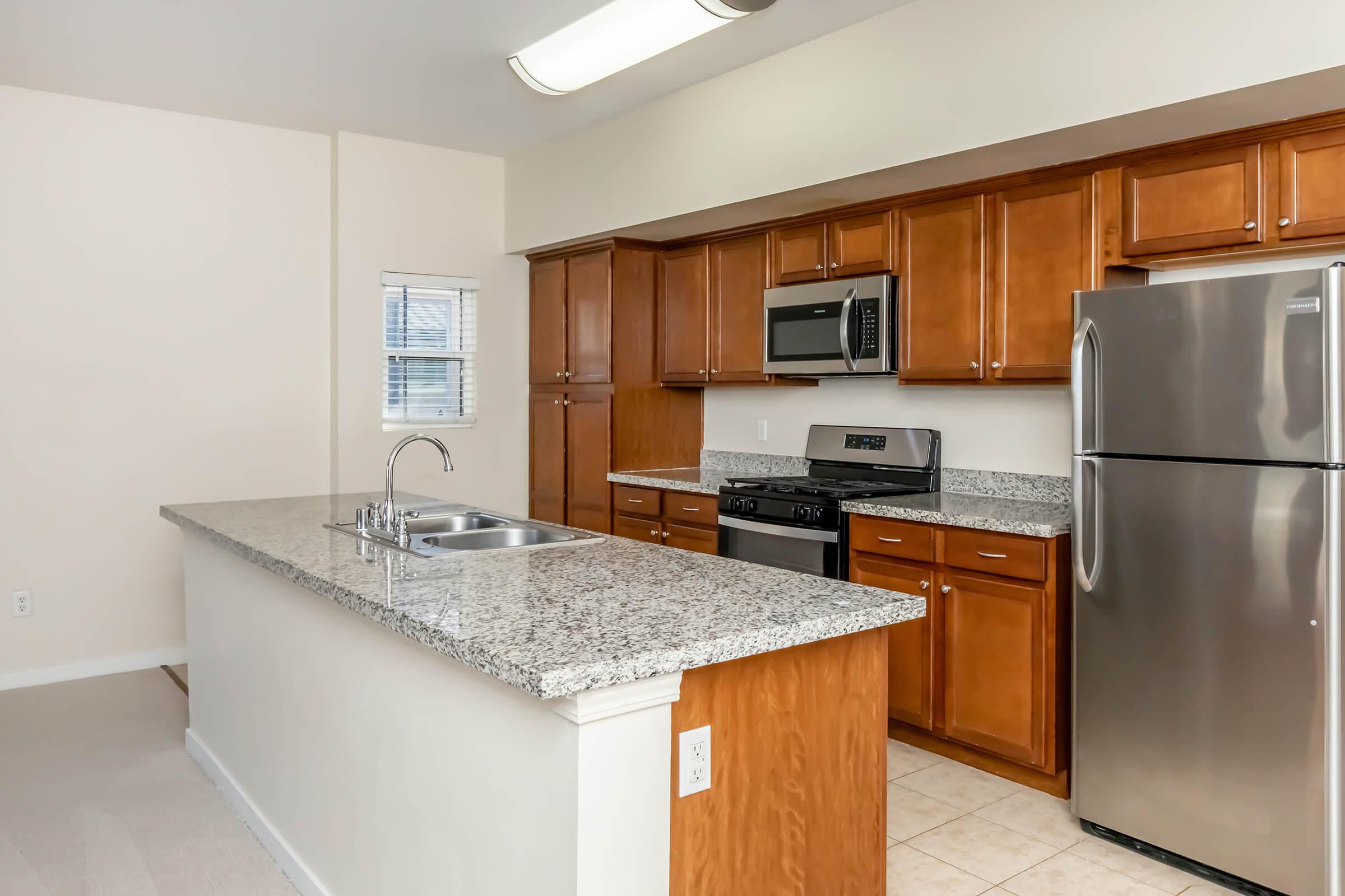 Modern kitchen featuring wooden cabinetry, granite countertop, stainless steel appliances including a microwave and refrigerator, and a double sink. The layout includes an island with additional counter space, and beige tile flooring complements the light-colored walls. Natural light streams in from a window.