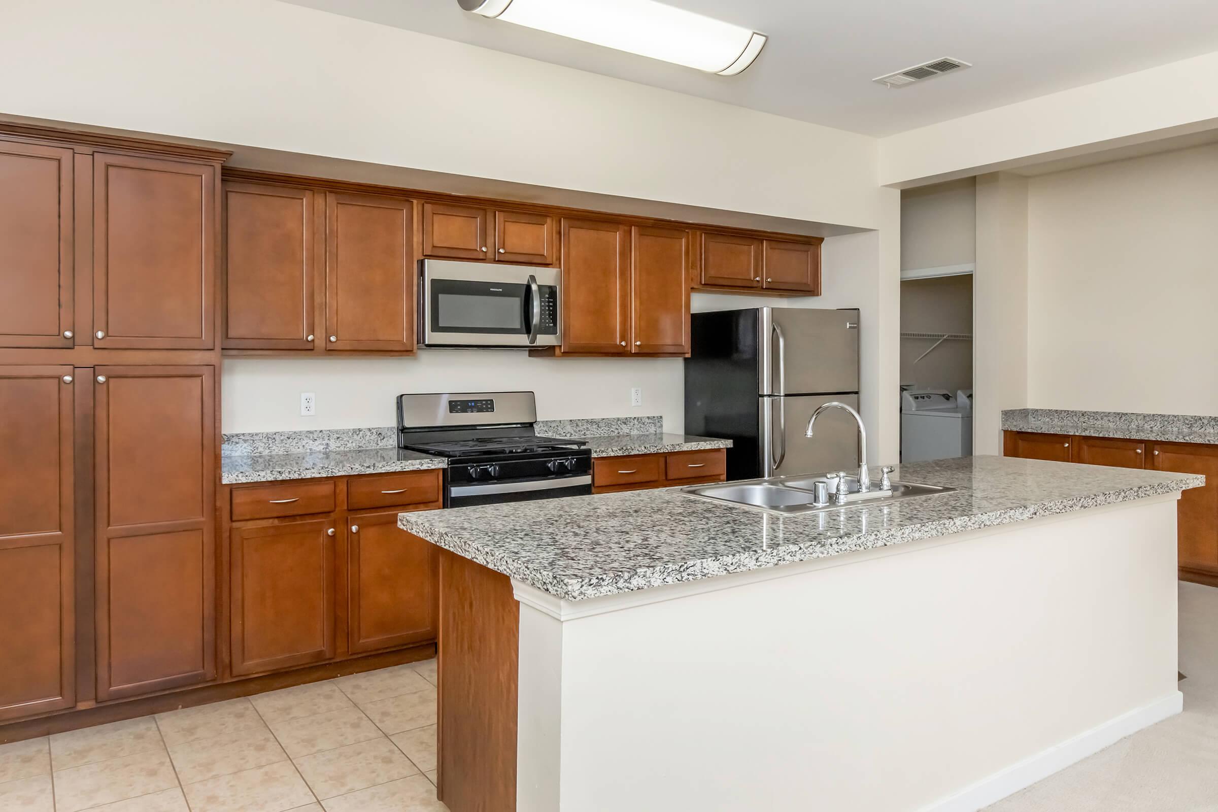 Modern kitchen with dark wood cabinets, granite countertops, and stainless steel appliances, including a microwave, stove, and refrigerator. The kitchen features a center island with a sink and has neutral-colored walls and tiled flooring. A laundry area is visible to the side.