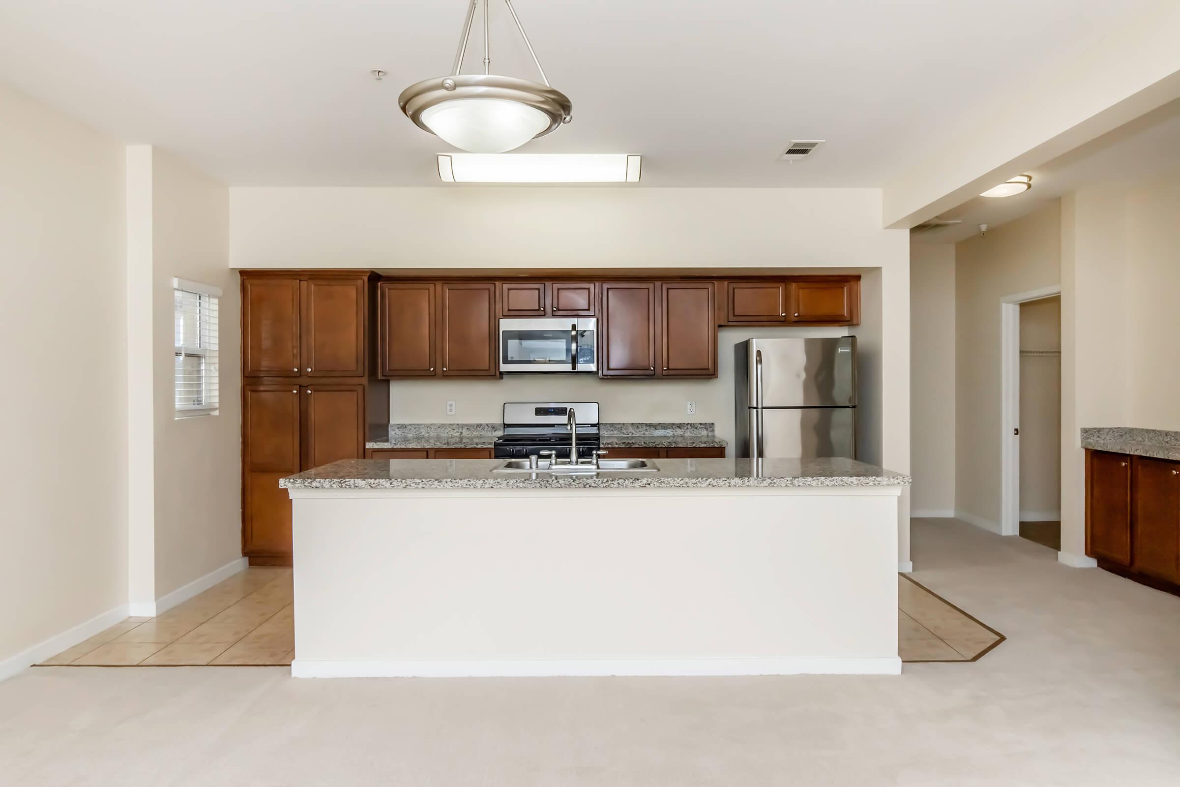Modern kitchen featuring wood cabinetry, granite countertops, and stainless steel appliances. The layout includes a central island with a sink, an open design leading to a spacious living area, beige walls, and plush carpet flooring. Natural light comes through windows, creating a welcoming atmosphere.