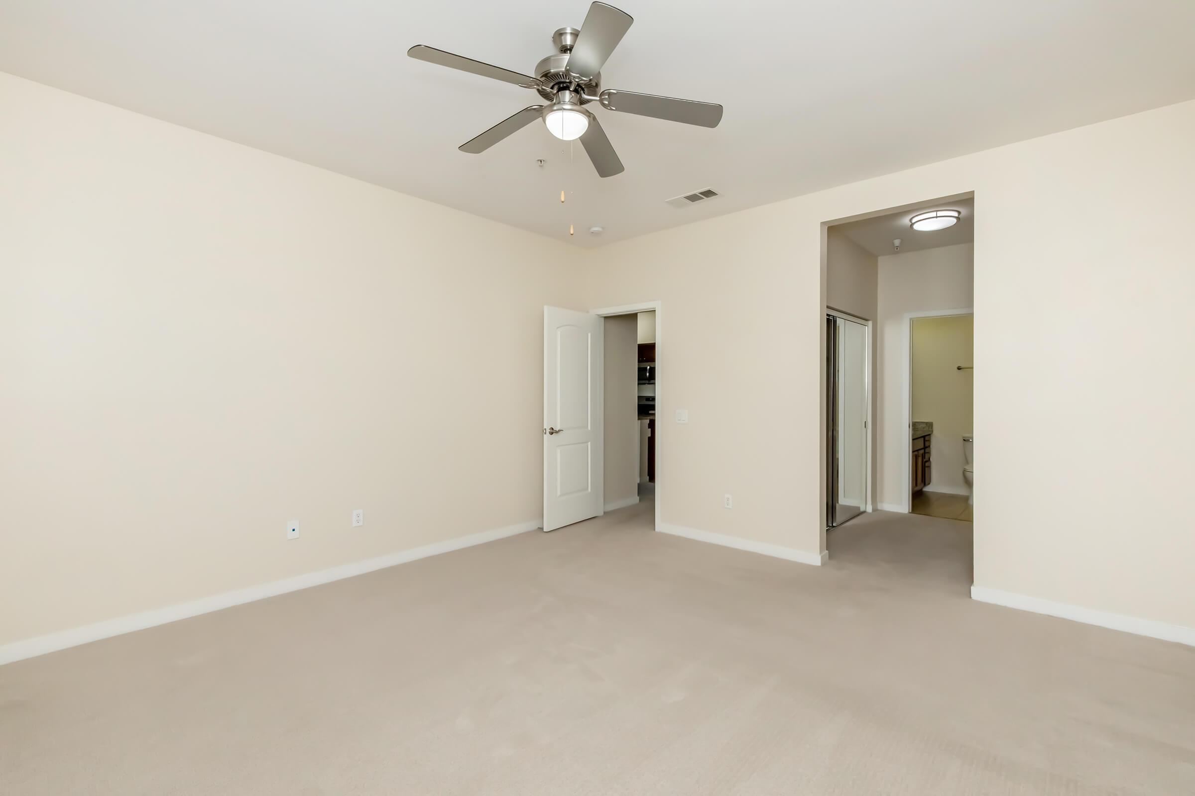 A spacious and empty room featuring beige walls and a light-colored carpet. A ceiling fan is mounted in the center. There is a doorway on the left leading to another room, and to the right, a glimpse of a bathroom can be seen. The overall atmosphere is bright and neutral.