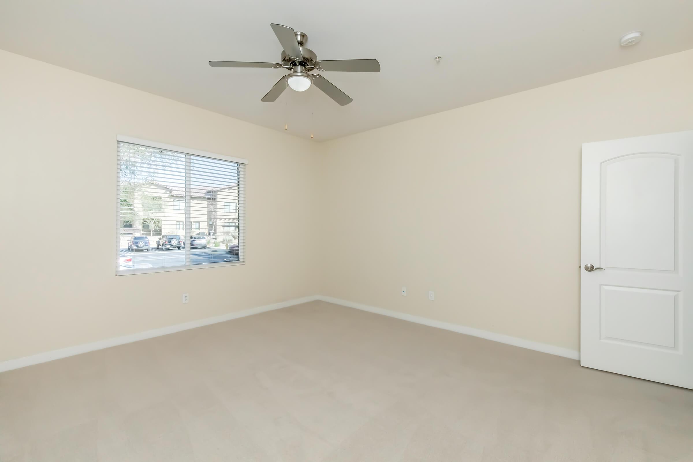 A spacious, empty room with beige walls and light brown carpet. It features a ceiling fan with three lights and a large window allowing natural light in. The window has white blinds and offers a view of a landscaped area outside. A white door is visible on the right side, leading to another part of the space.