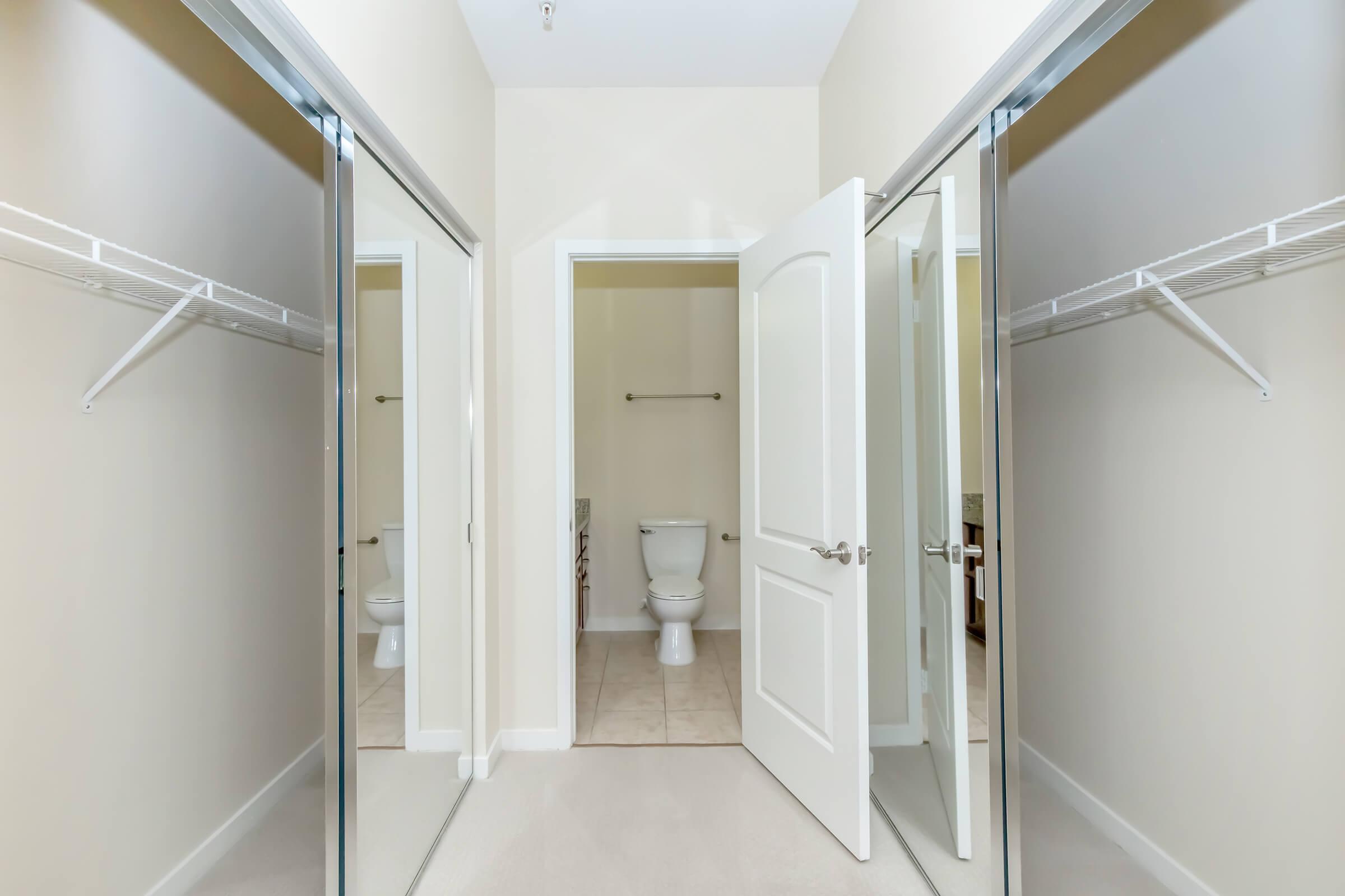 A spacious room featuring two mirrored sliding closet doors. In the center, a white door leads to a bathroom with a toilet visible. Light beige walls and tile flooring create a bright, clean atmosphere. The room has a simple, modern design with ample storage space.
