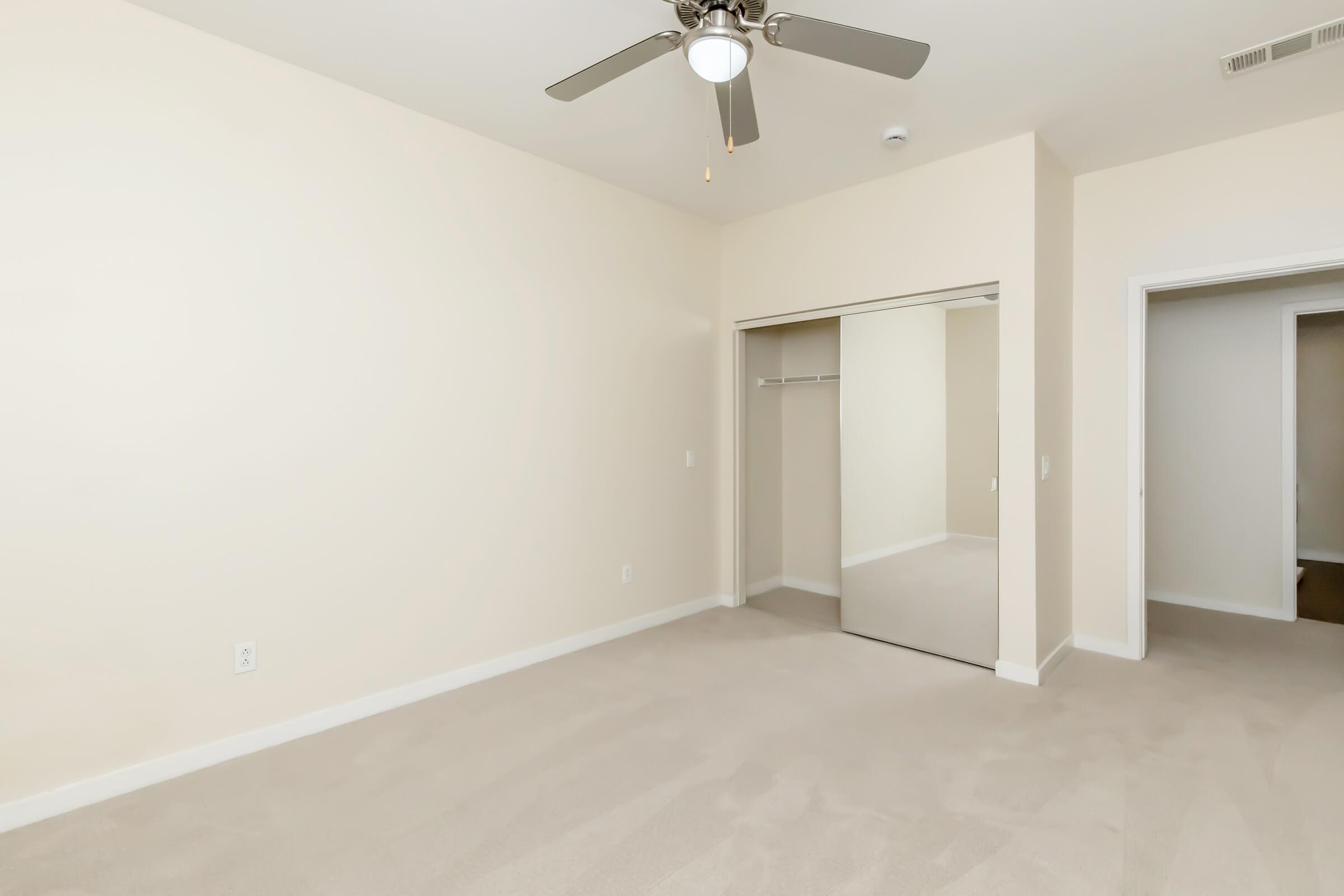 A spacious, empty bedroom featuring light-colored walls and carpet. A ceiling fan is visible, along with a mirrored closet door on the left. The room has one open doorway leading to another area, providing a bright and airy feel. Ideal for personalization and design.