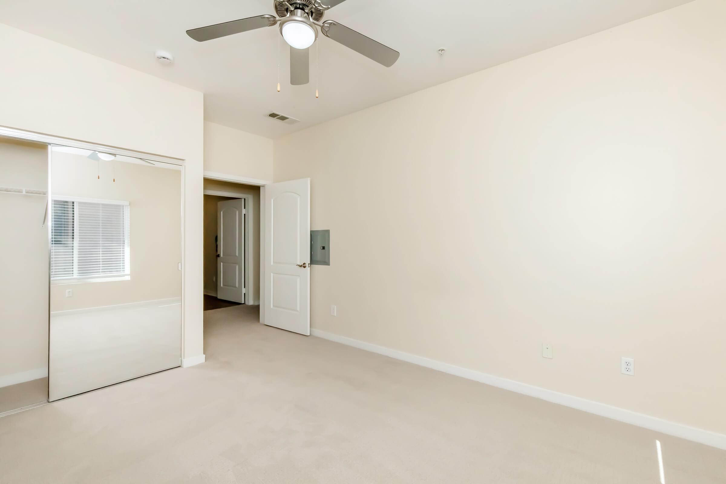 A well-lit room with beige walls and carpet, featuring a ceiling fan. There is a large mirror on the left wall and a partially open door leading to another room. Natural light enters through a window, creating a bright and spacious atmosphere.